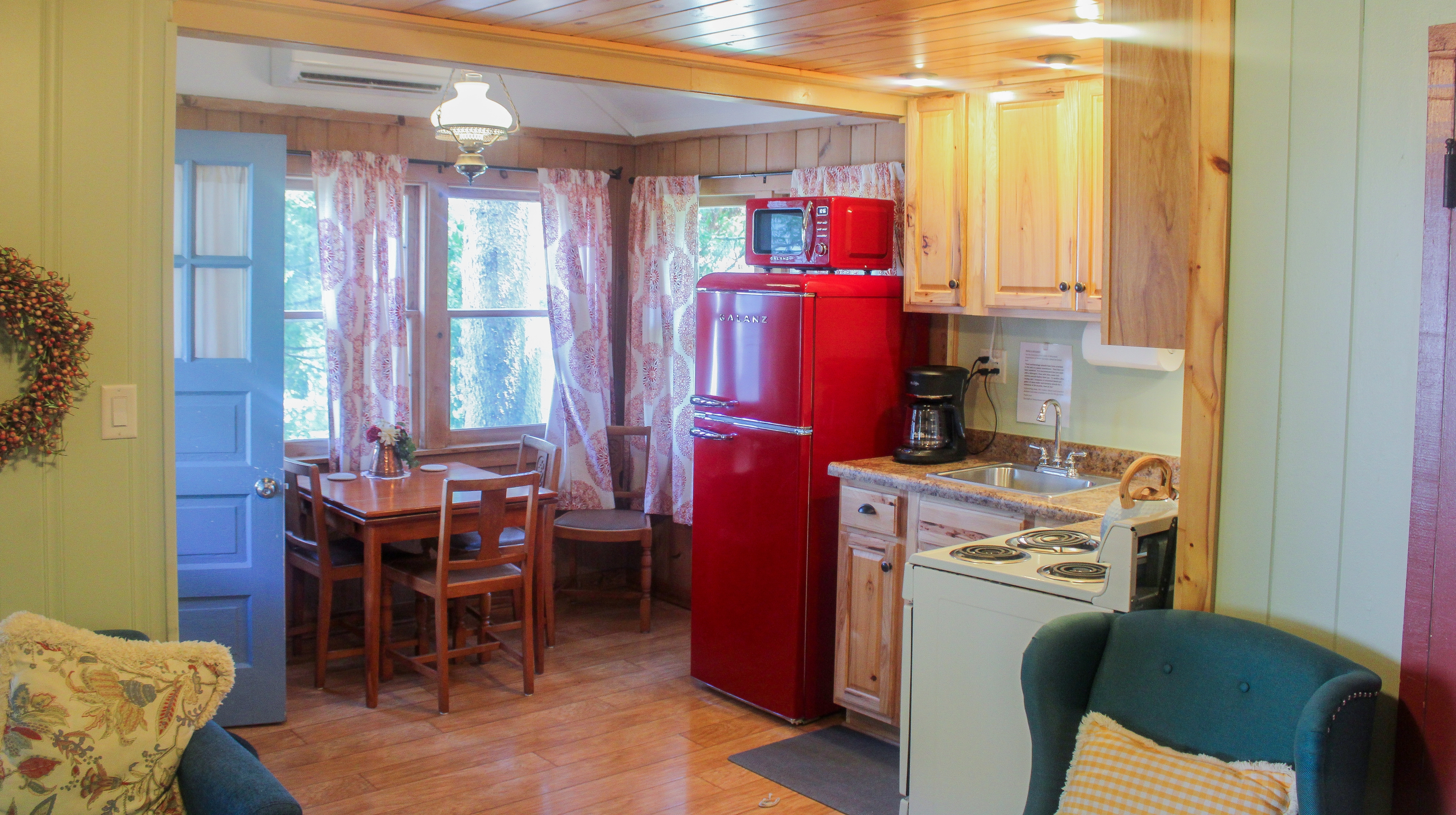 A rustic cottage interior featuring a small kitchen with wooden cabinets, a dining area with a wooden table, and warm lighting.