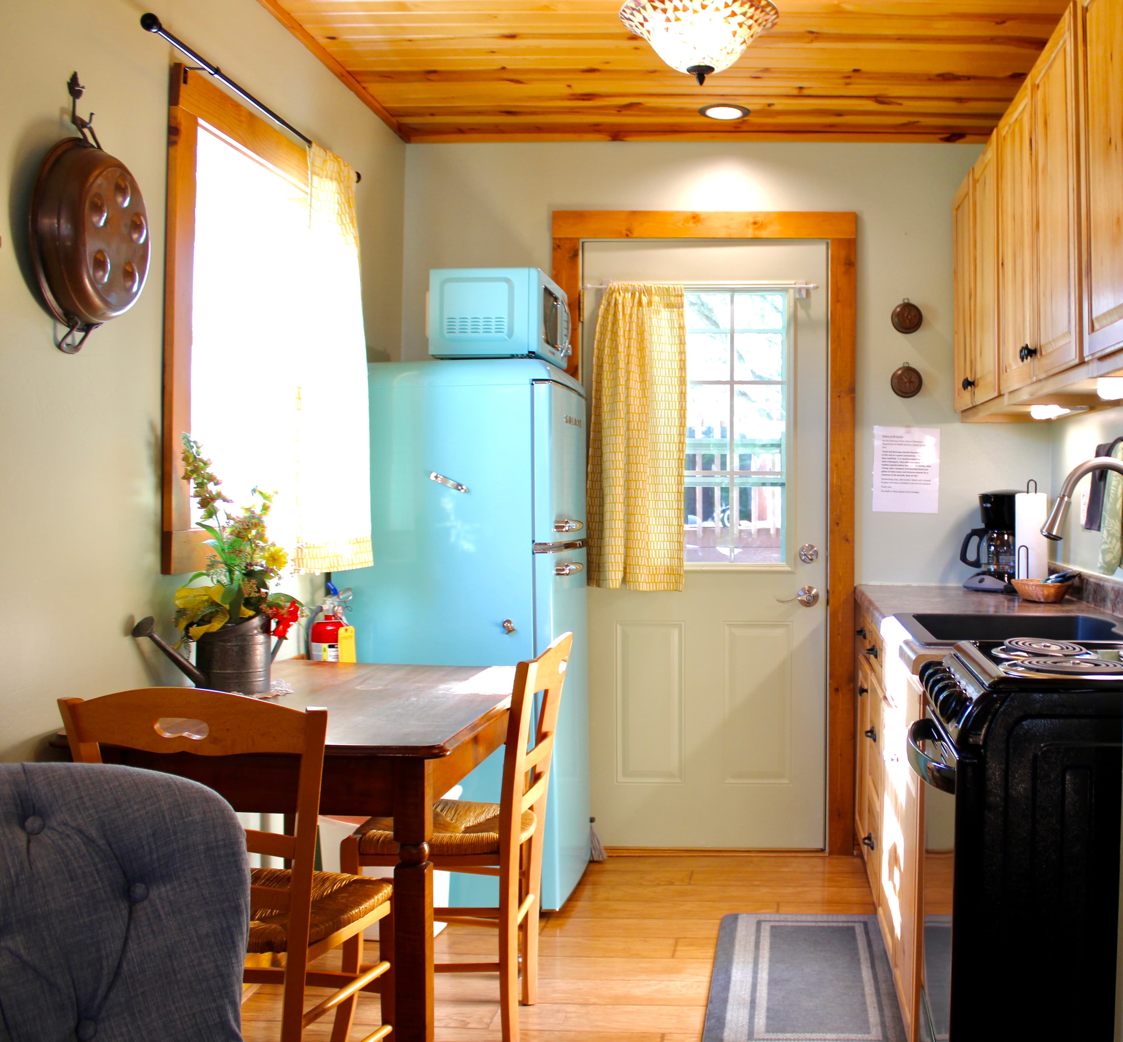 A small, functional kitchen with light wood cabinets, a white stove, a dining table for two, and a bright yellow curtain on the window.
