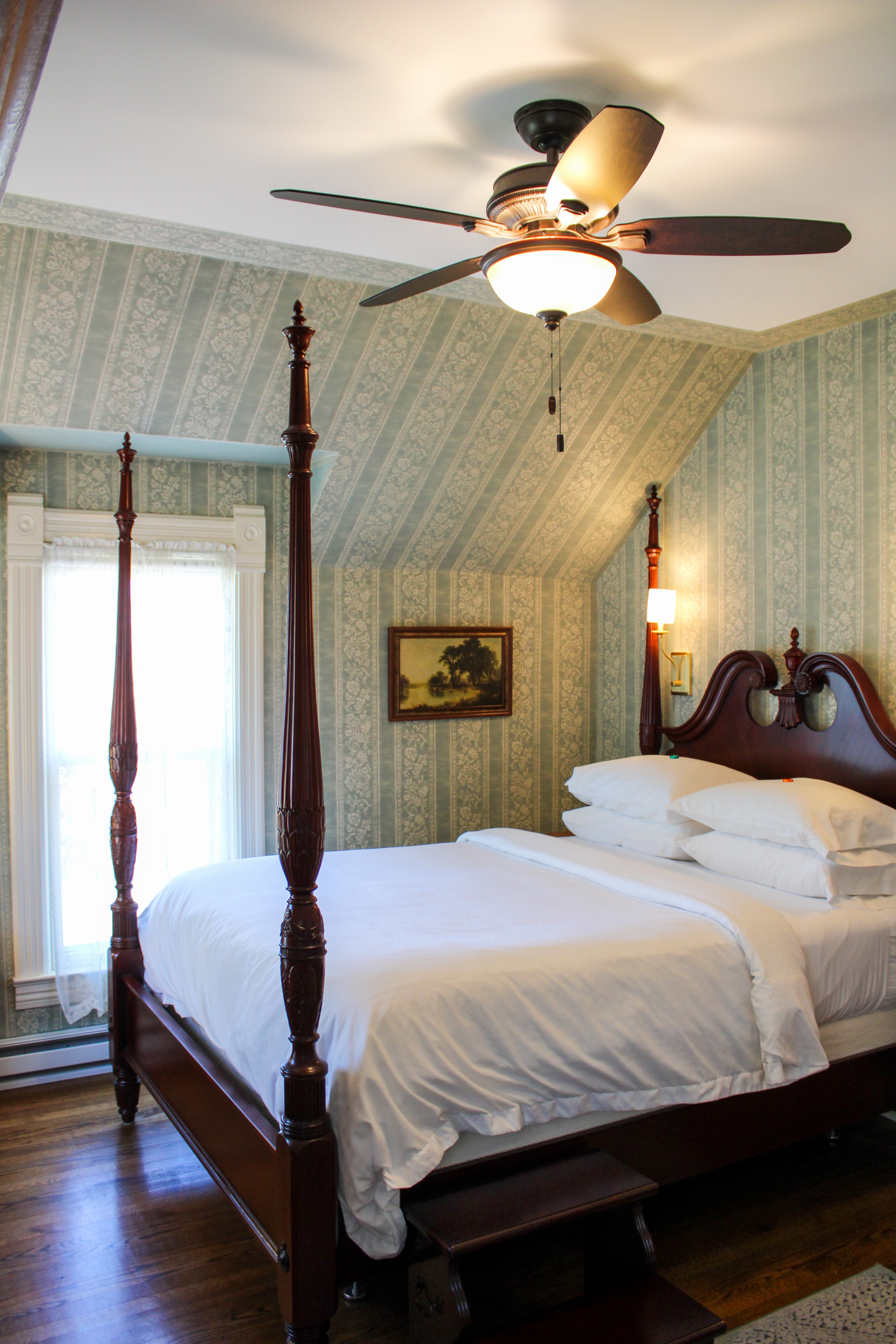 A bright bedroom with a wooden four-poster bed, patterned wallpaper, a television on a dresser, and a window letting in sunlight.