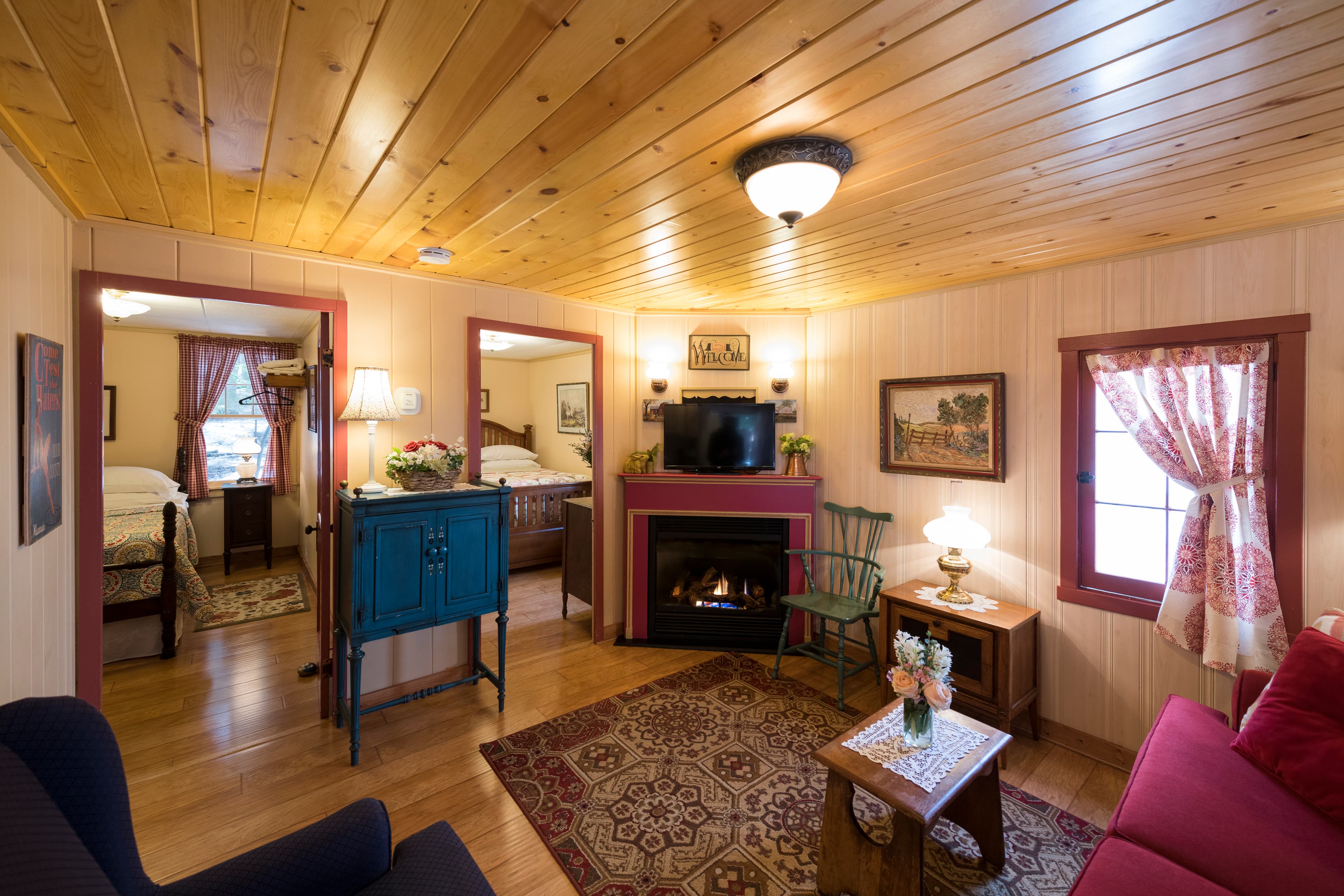A charming cottage interior with wood-paneled ceilings, a cozy living room with a fireplace, and views into two adjoining bedrooms.