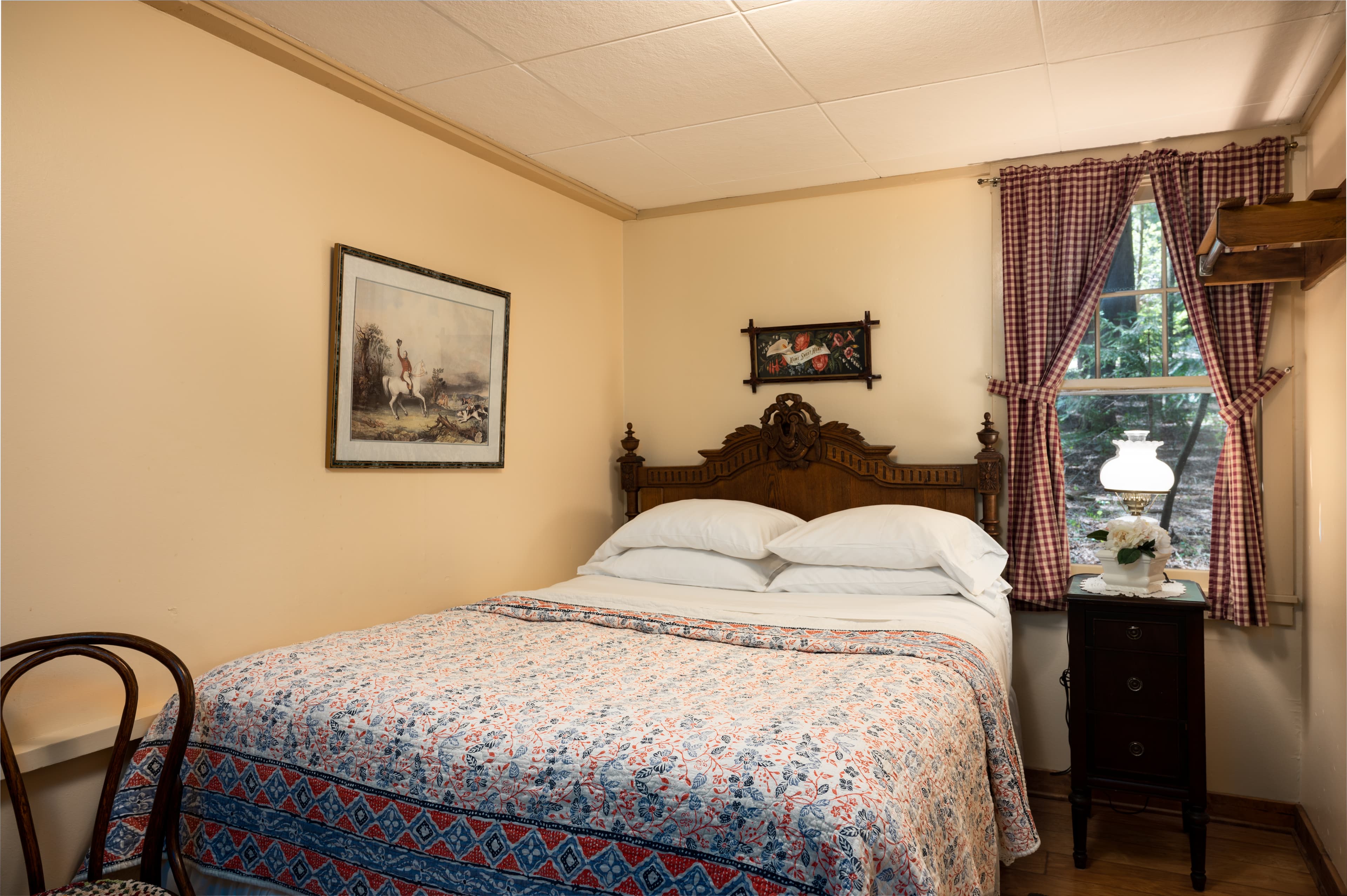 A charming bedroom with light peach walls, a wooden bed with a colorful quilt, a window with purple curtains, and a classic lamp.