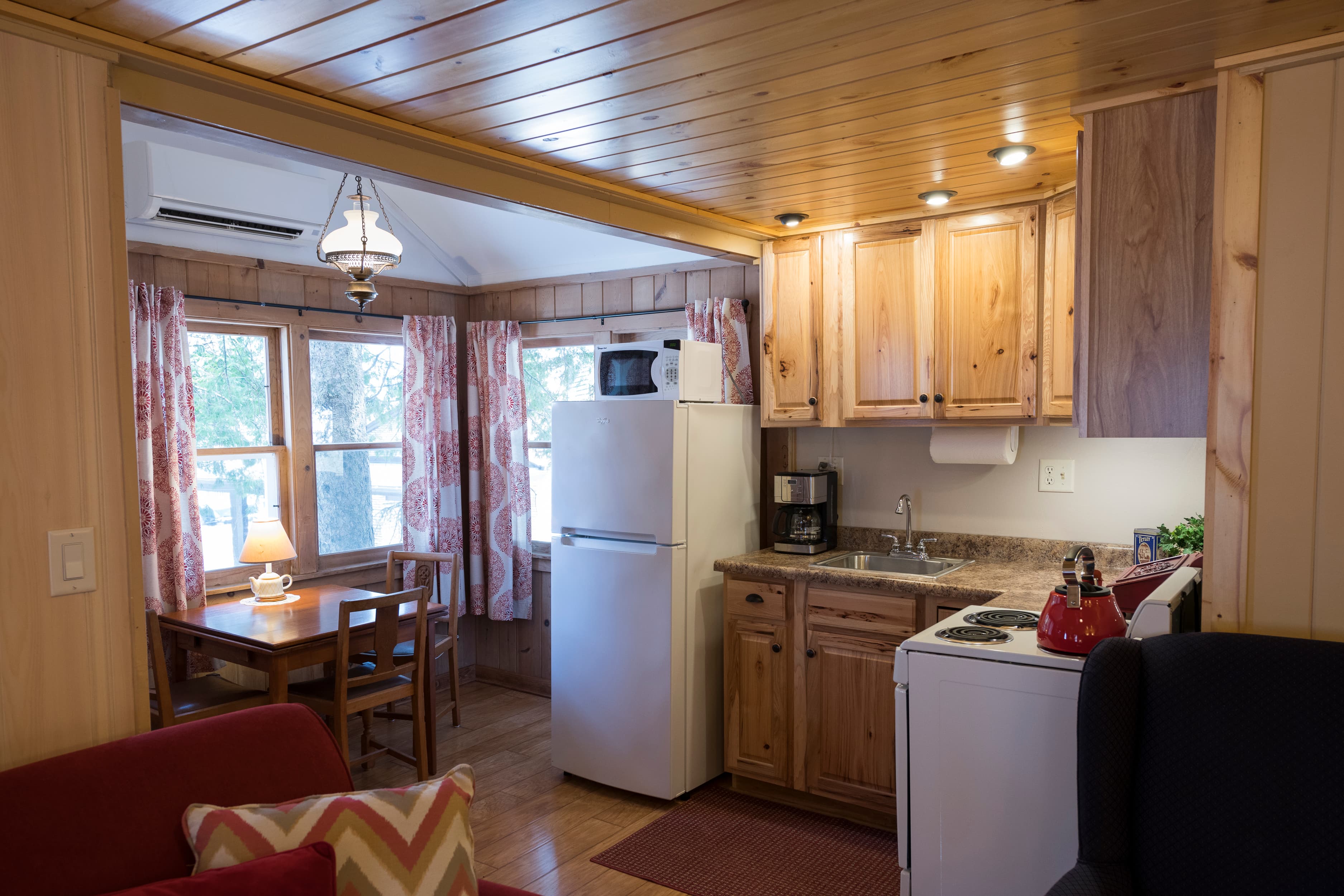 A rustic cottage interior featuring a small kitchen with wooden cabinets, a dining area with a wooden table, and warm lighting.