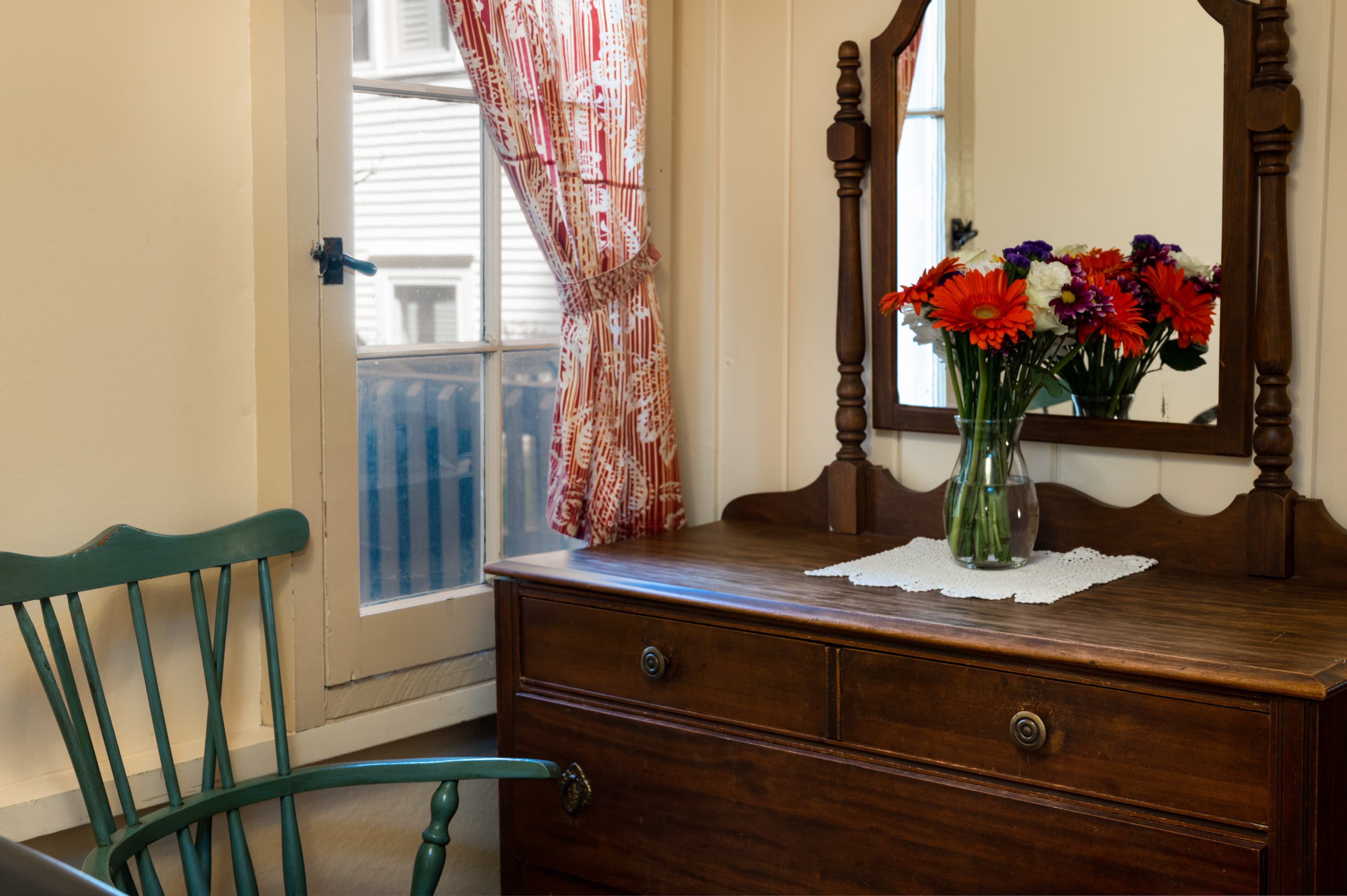 A charming wooden dresser with an ornate mirror and a vase of colorful flowers, next to a window with patterned curtains and a green chair.