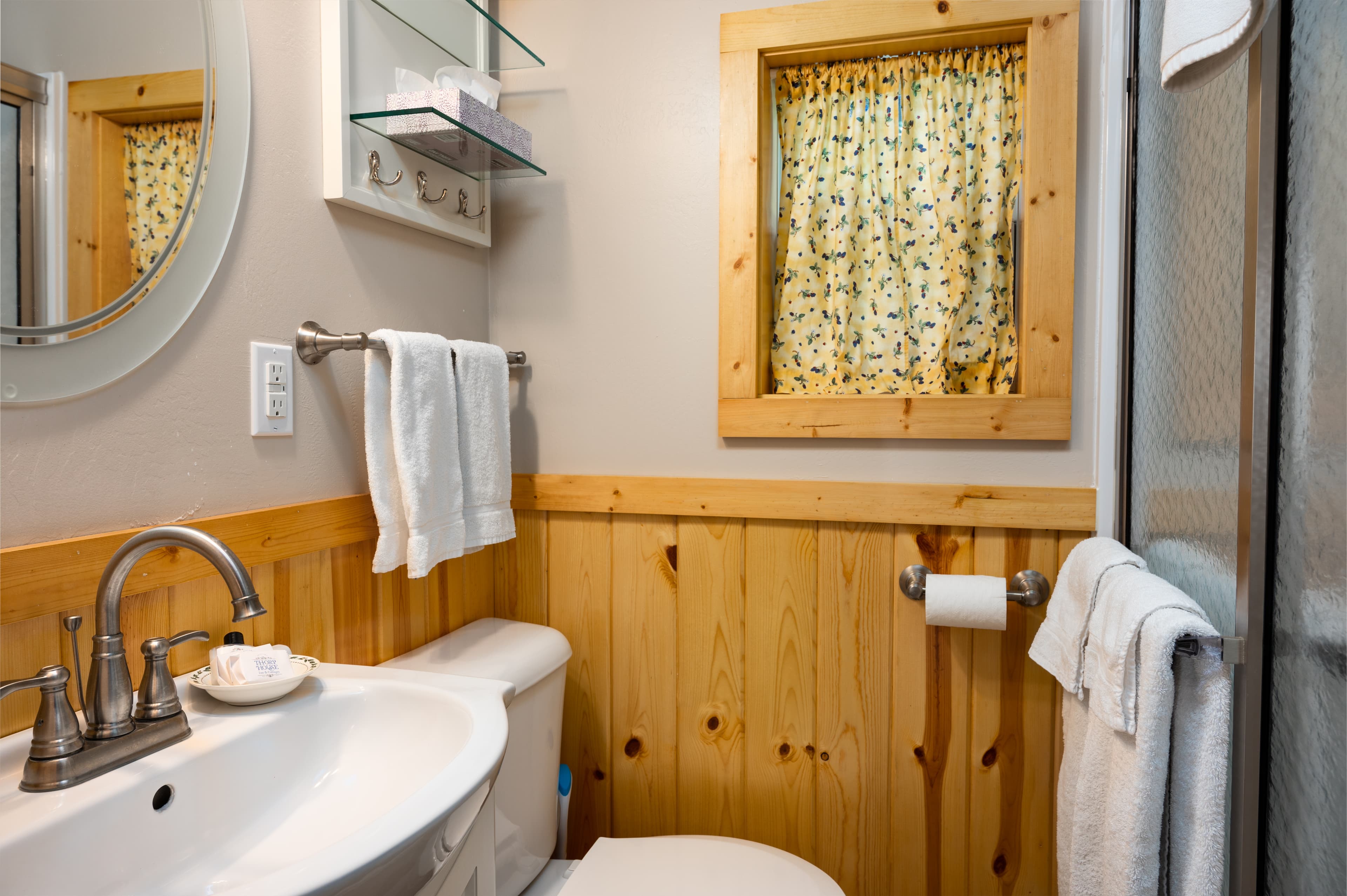A well-appointed bathroom with a white sink, toilet, and wood paneling, featuring a window with a patterned curtain and a glass shower.