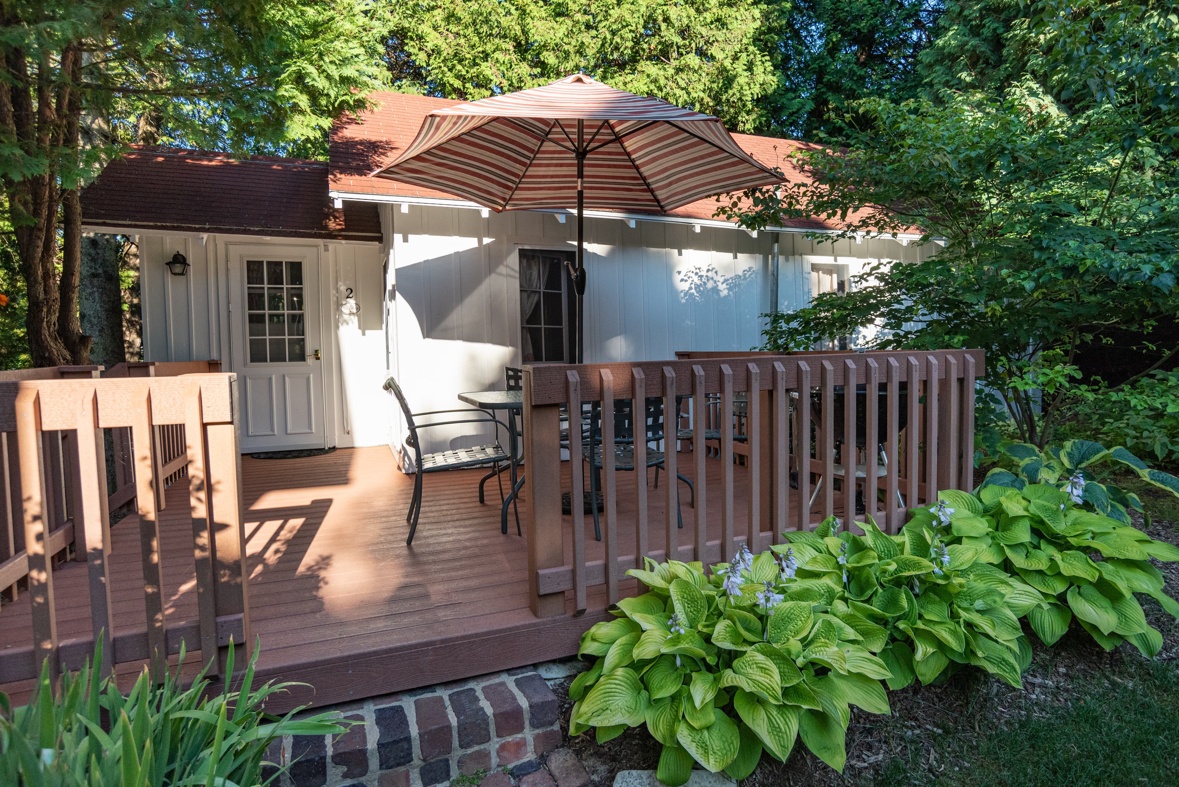 A charming cottage with a red roof, a spacious wooden deck with an umbrella and outdoor seating, surrounded by lush greenery.