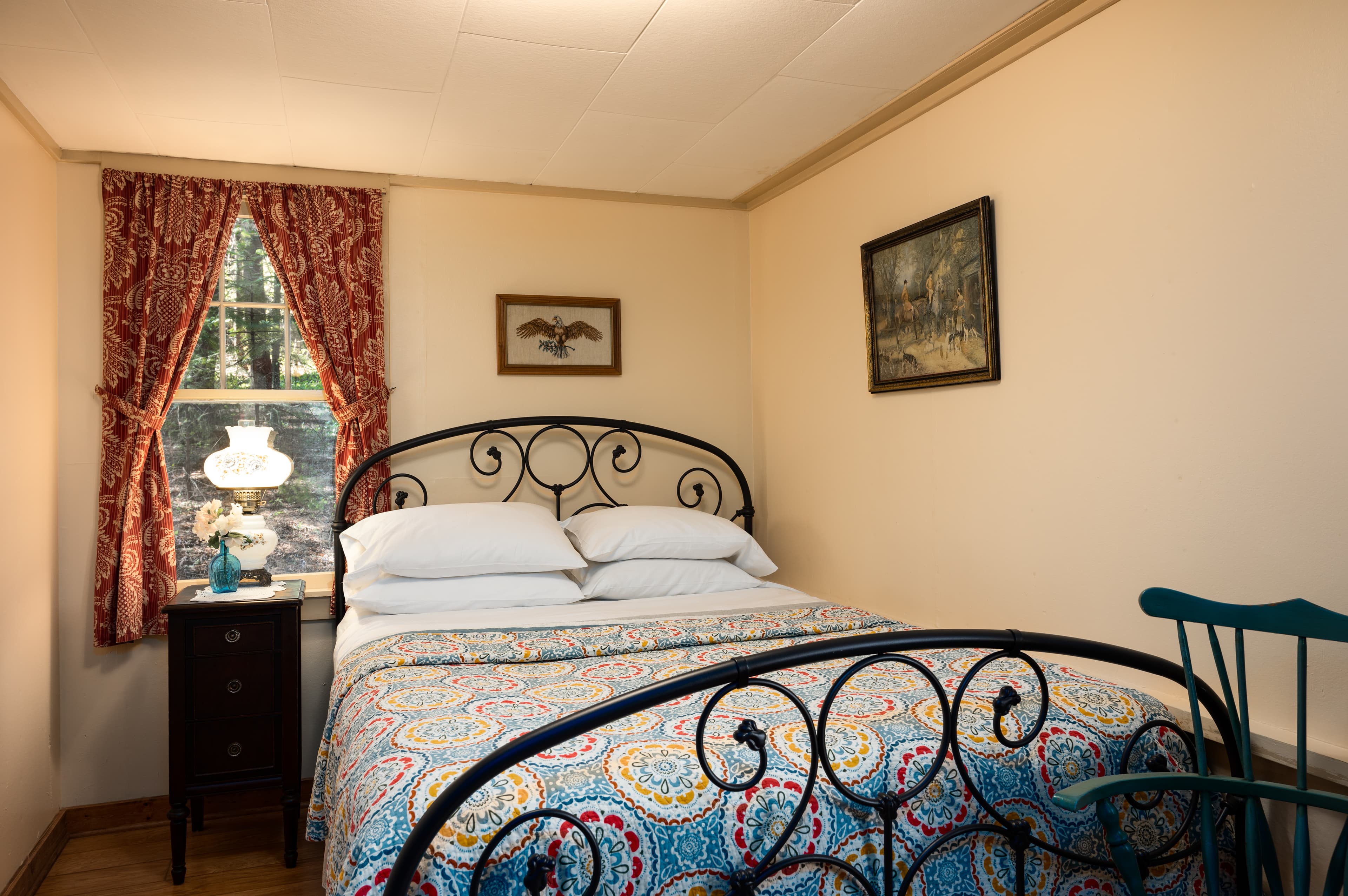 A quaint bedroom featuring a wrought iron bed with a colorful quilt, light-colored walls, and a window with red patterned curtains.