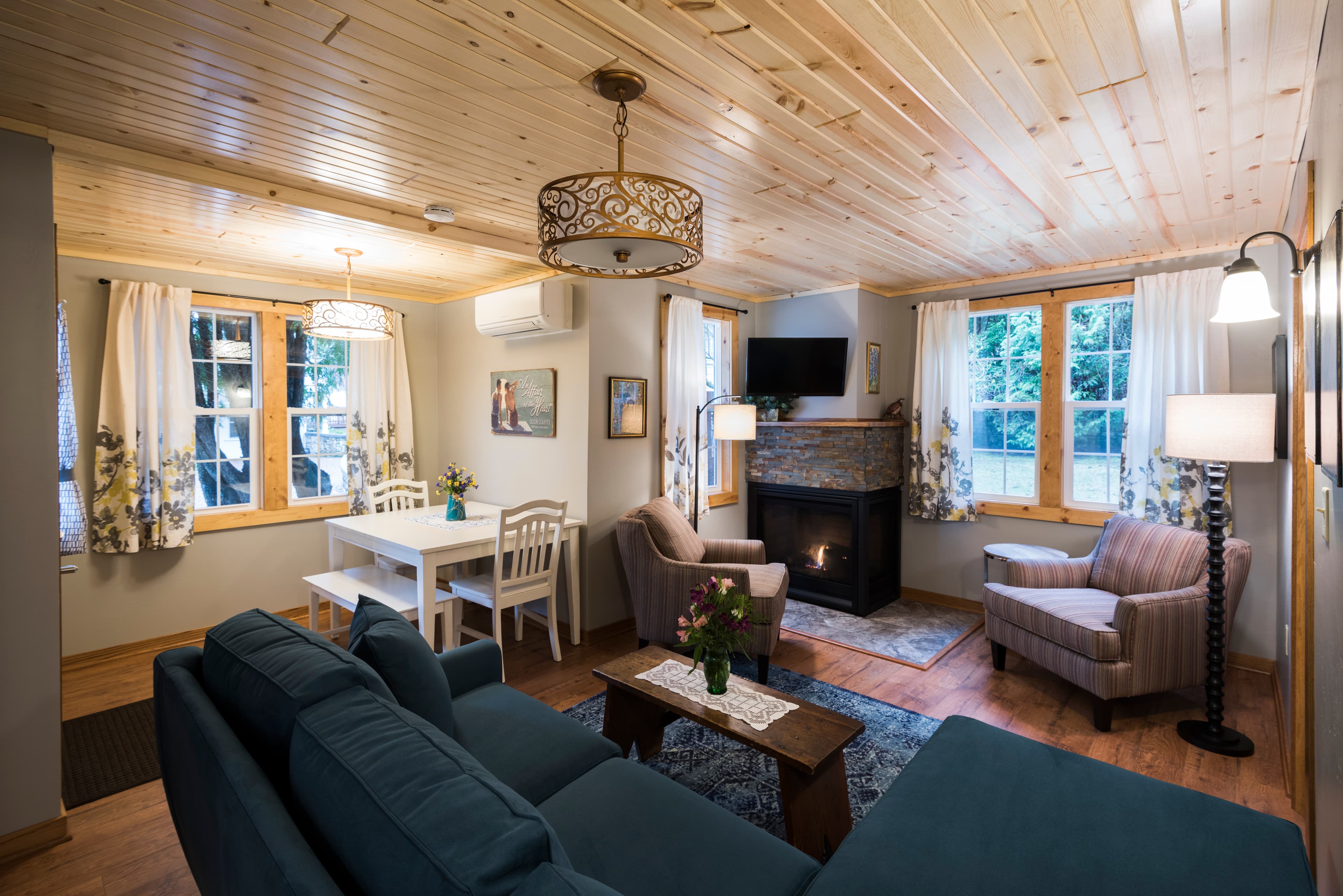 A spacious and inviting living area with a comfortable blue sofa, a stone fireplace, a dining table, and warm wood-paneled ceilings.