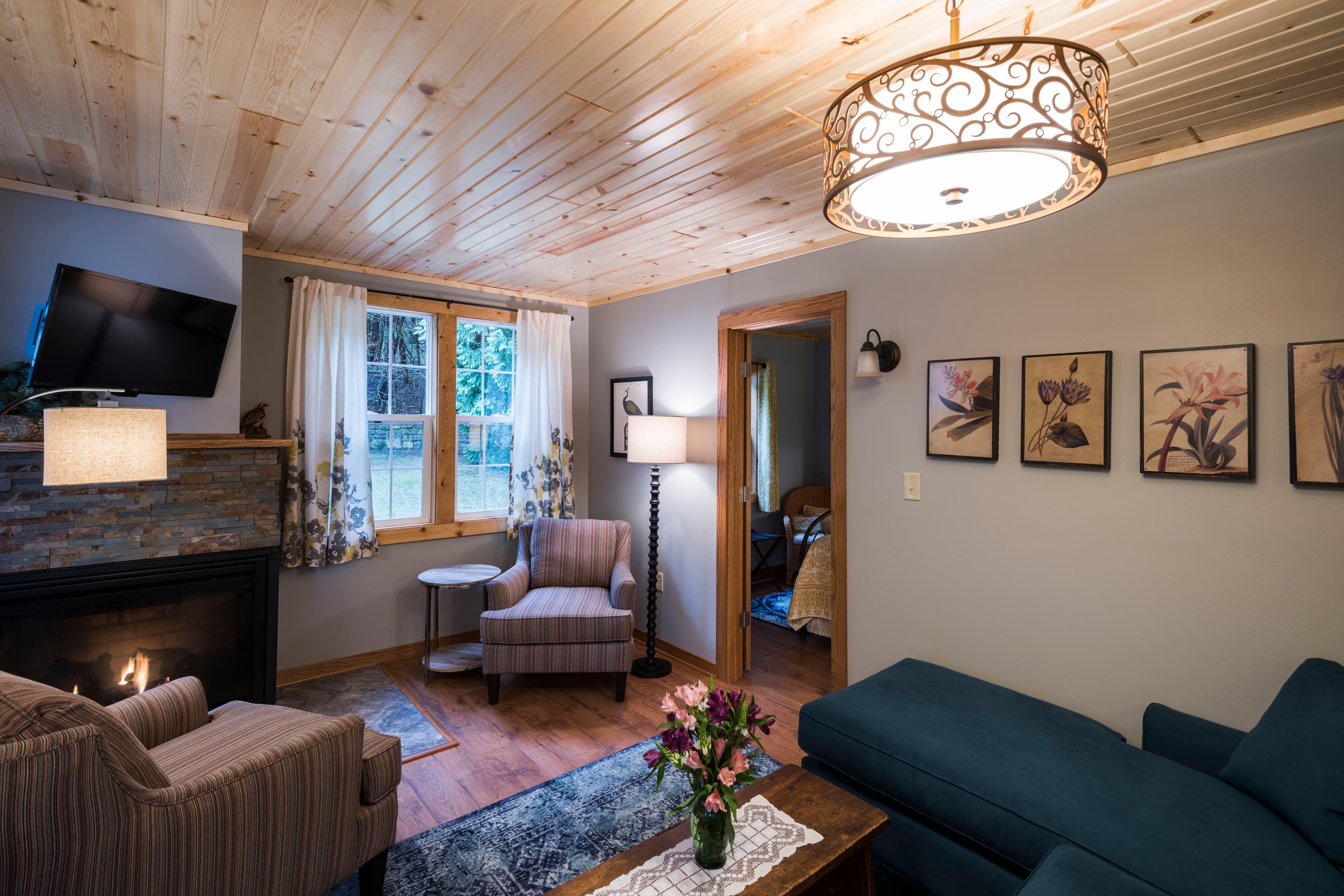 A comfortable living room with a fireplace, a large blue sofa, a patterned armchair, and wood-paneled ceilings, leading to a bedroom.