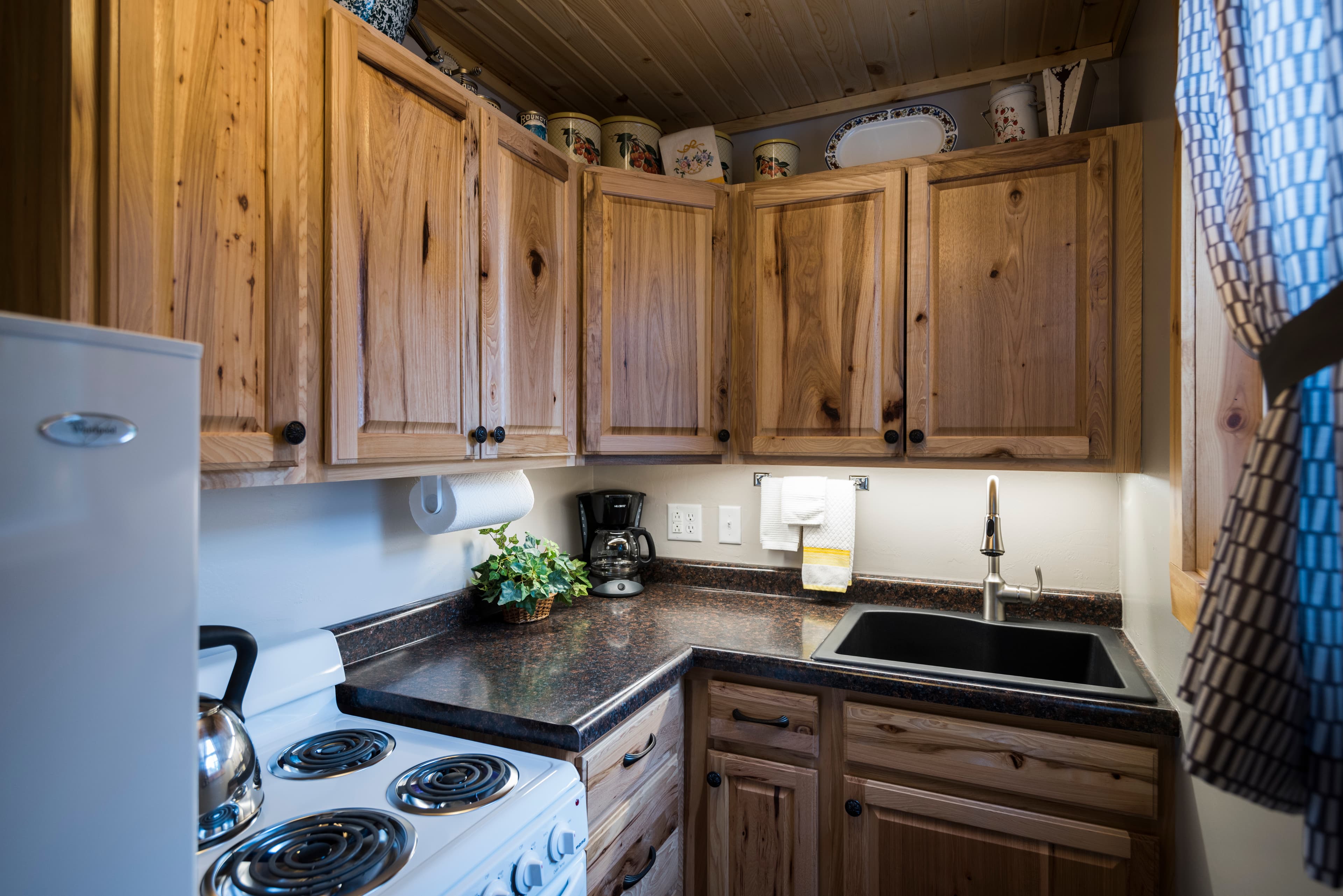 A compact kitchen featuring wooden cabinets, a black sink, a white stove, and a small refrigerator, with a wood-paneled ceiling.
