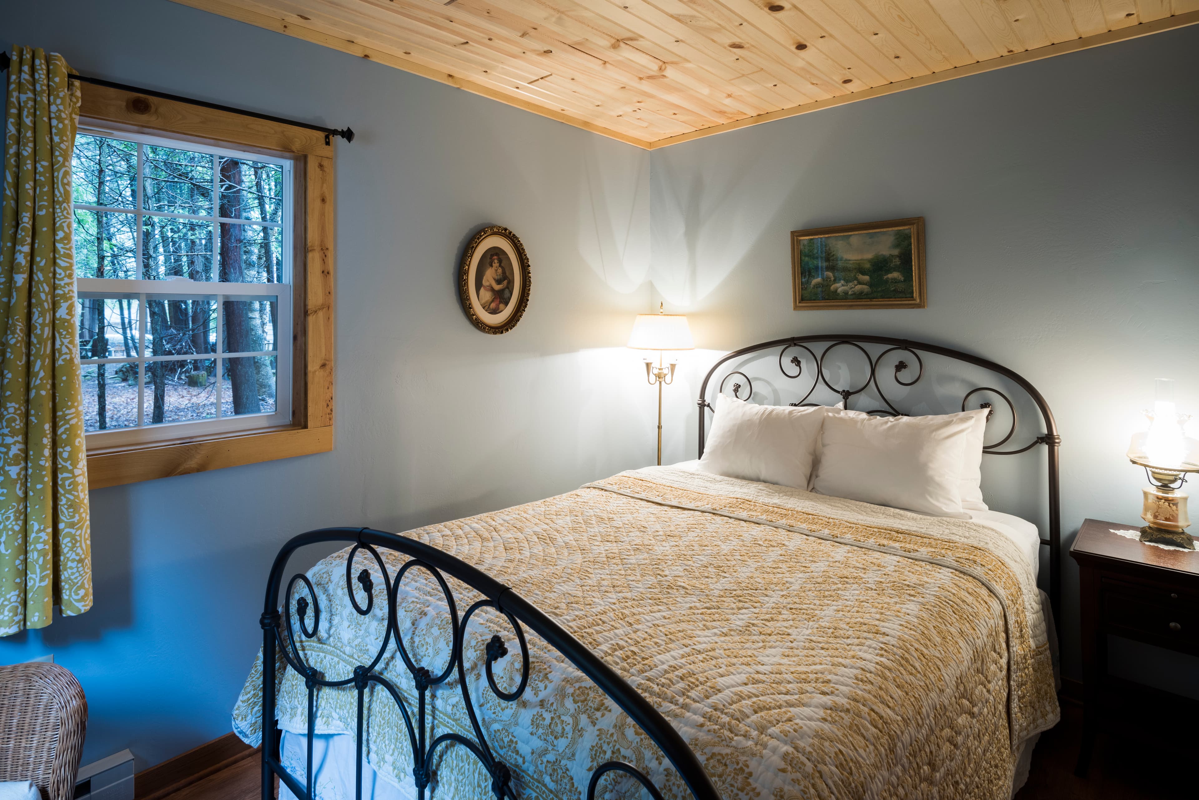 A tranquil bedroom featuring a wrought iron bed with a yellow patterned quilt, light blue walls, and a window offering a view of trees.