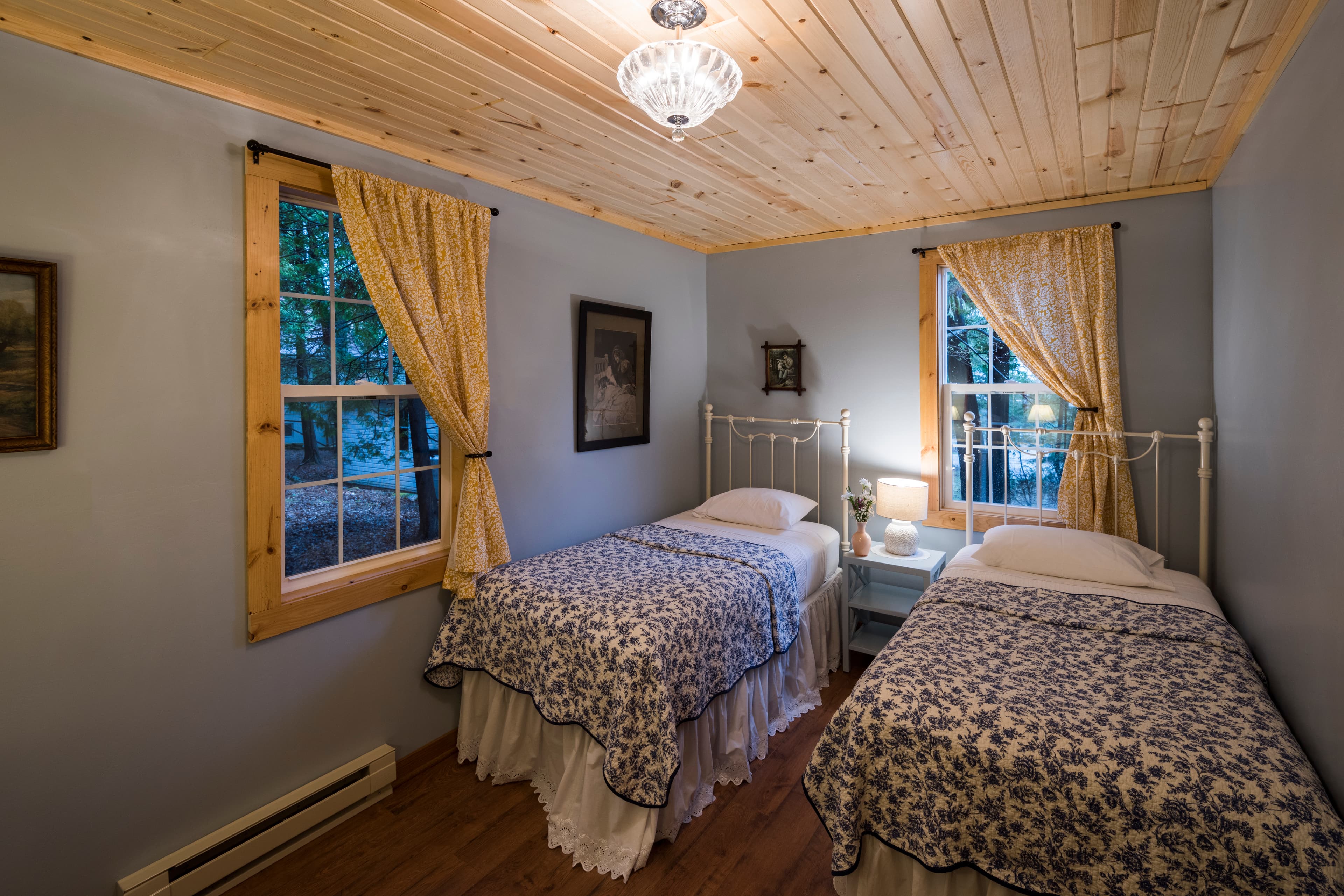 A charming bedroom featuring two twin beds with blue and white patterned quilts, light blue walls, and a warm wood-paneled ceiling.