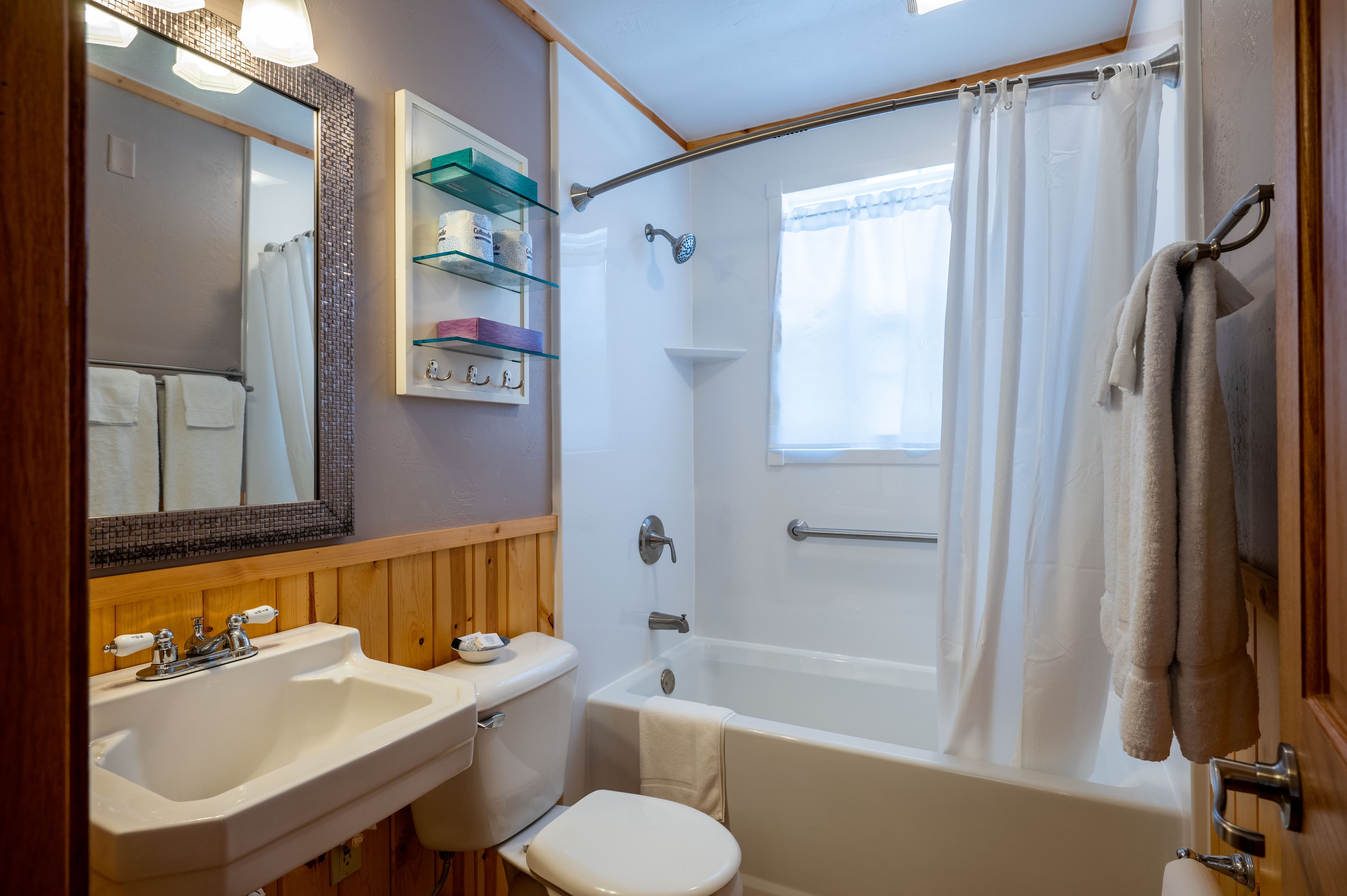 A rustic bathroom featuring light purple walls, wood paneling, a white sink and toilet, and a bathtub with a white shower curtain.