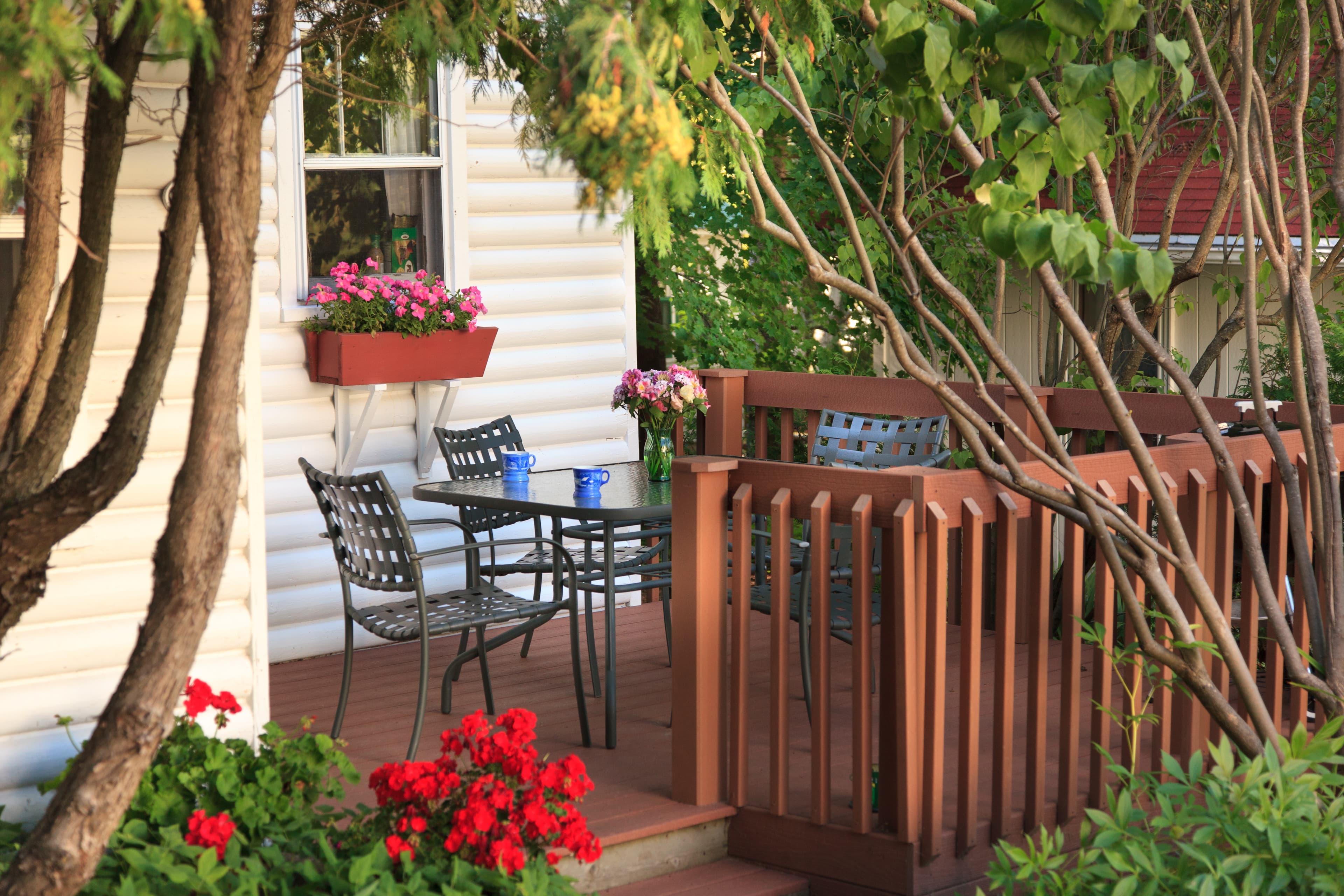 A welcoming outdoor deck with a table and chairs, adorned with vibrant red and pink flowers, next to a white cottage.