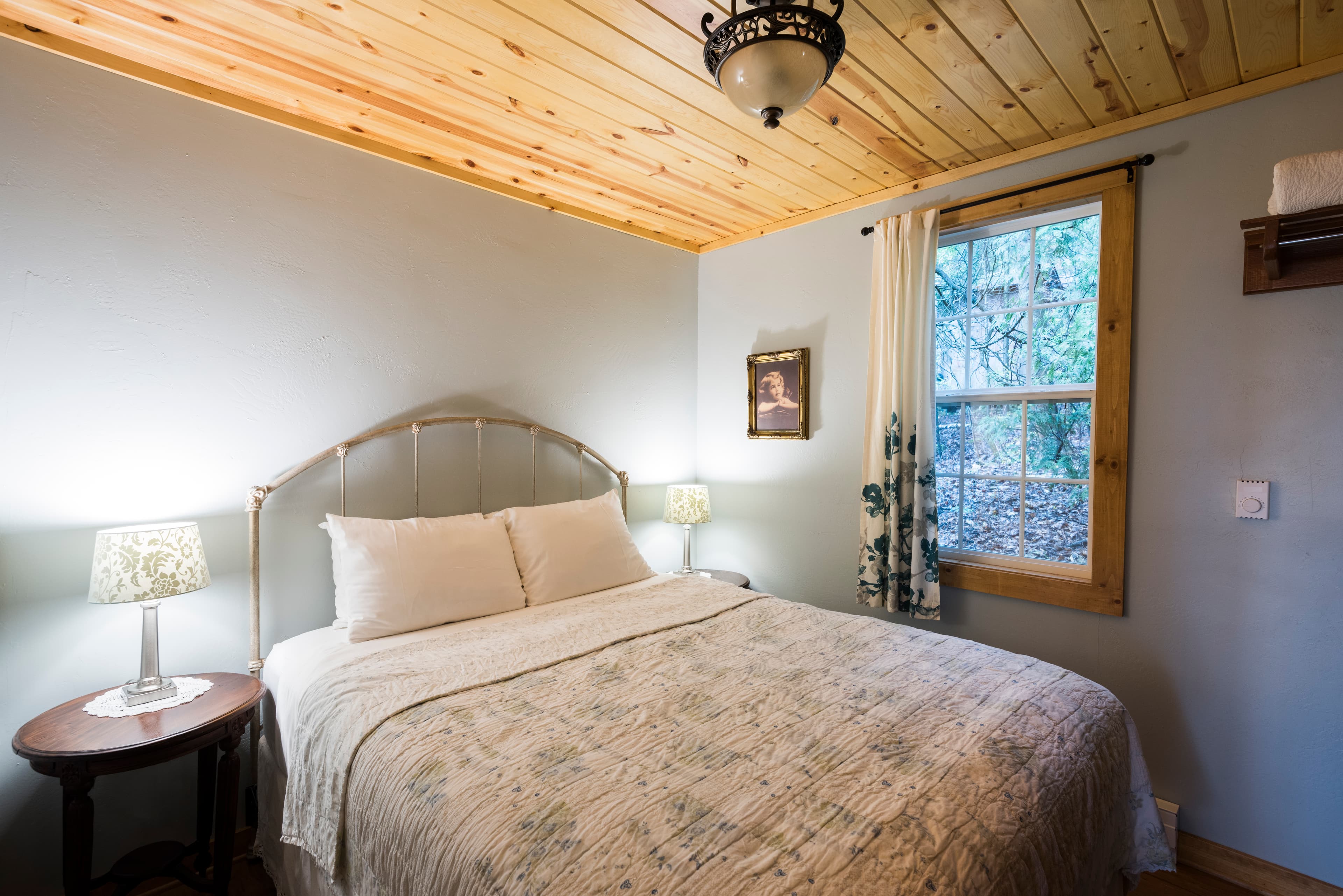 A tranquil bedroom featuring a bed with a light-colored headboard, pale gray walls, a rustic wood-paneled ceiling, and a window with patterned curtains.