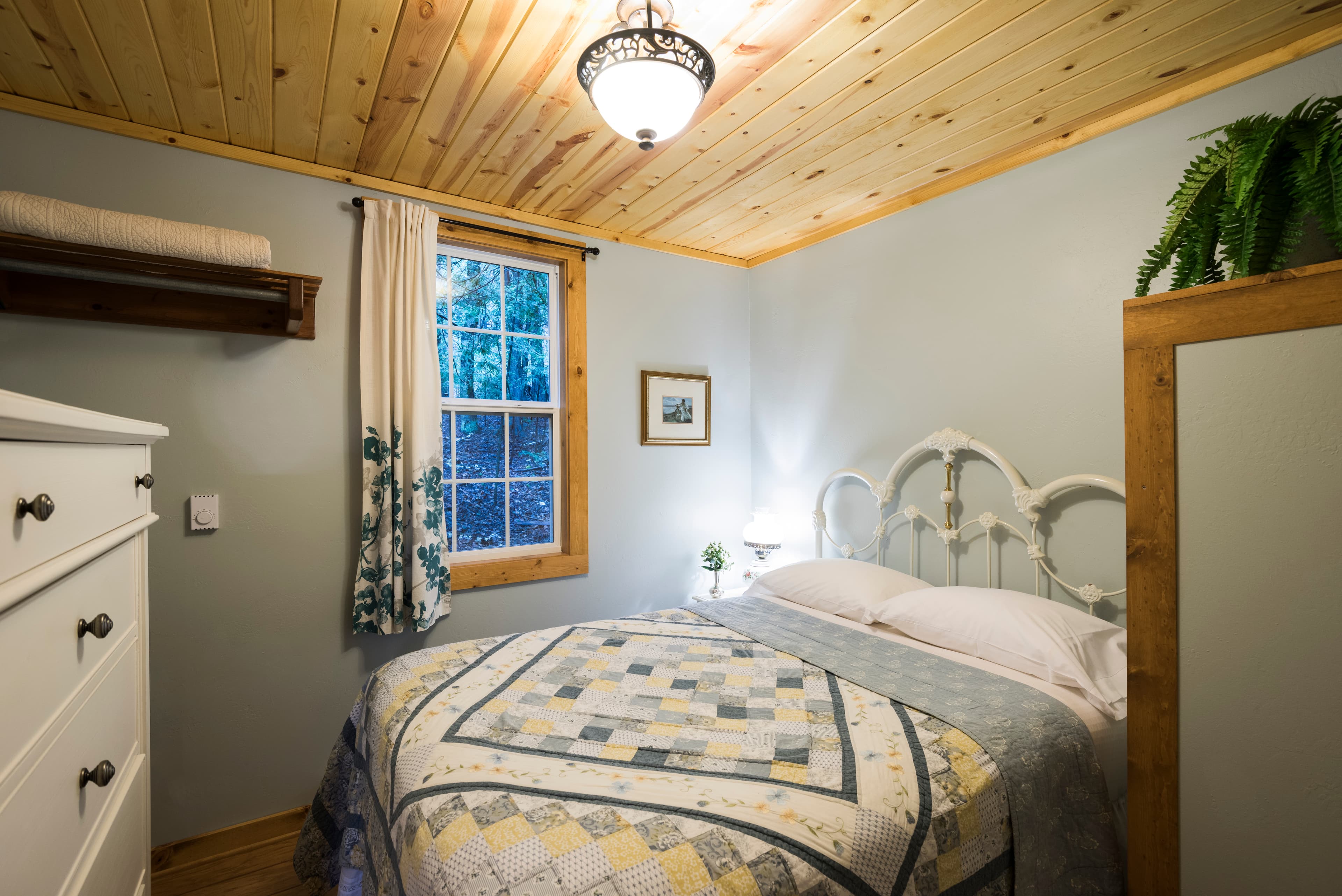 A tranquil bedroom featuring a white metal bed with a patterned quilt, light gray walls, and a rustic wood-paneled ceiling.