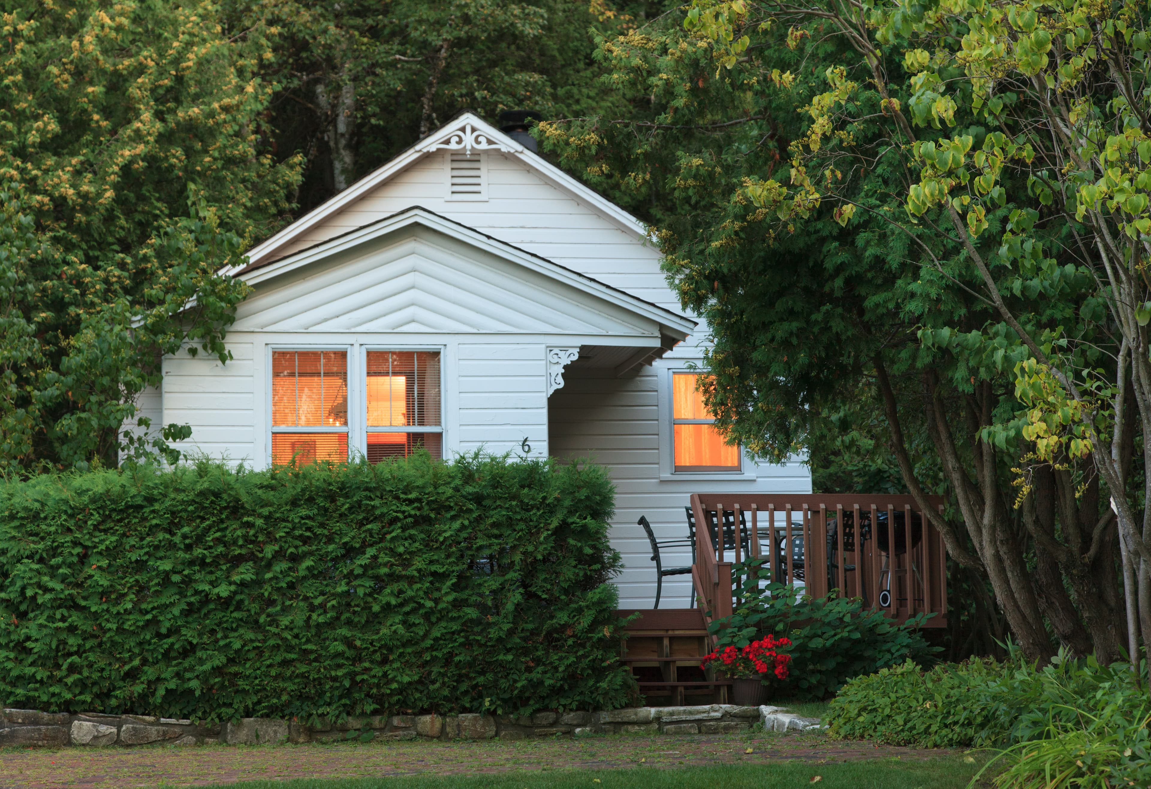A cozy white cottage with warm lights glowing from its windows, nestled among green trees and bushes, with a small wooden deck.