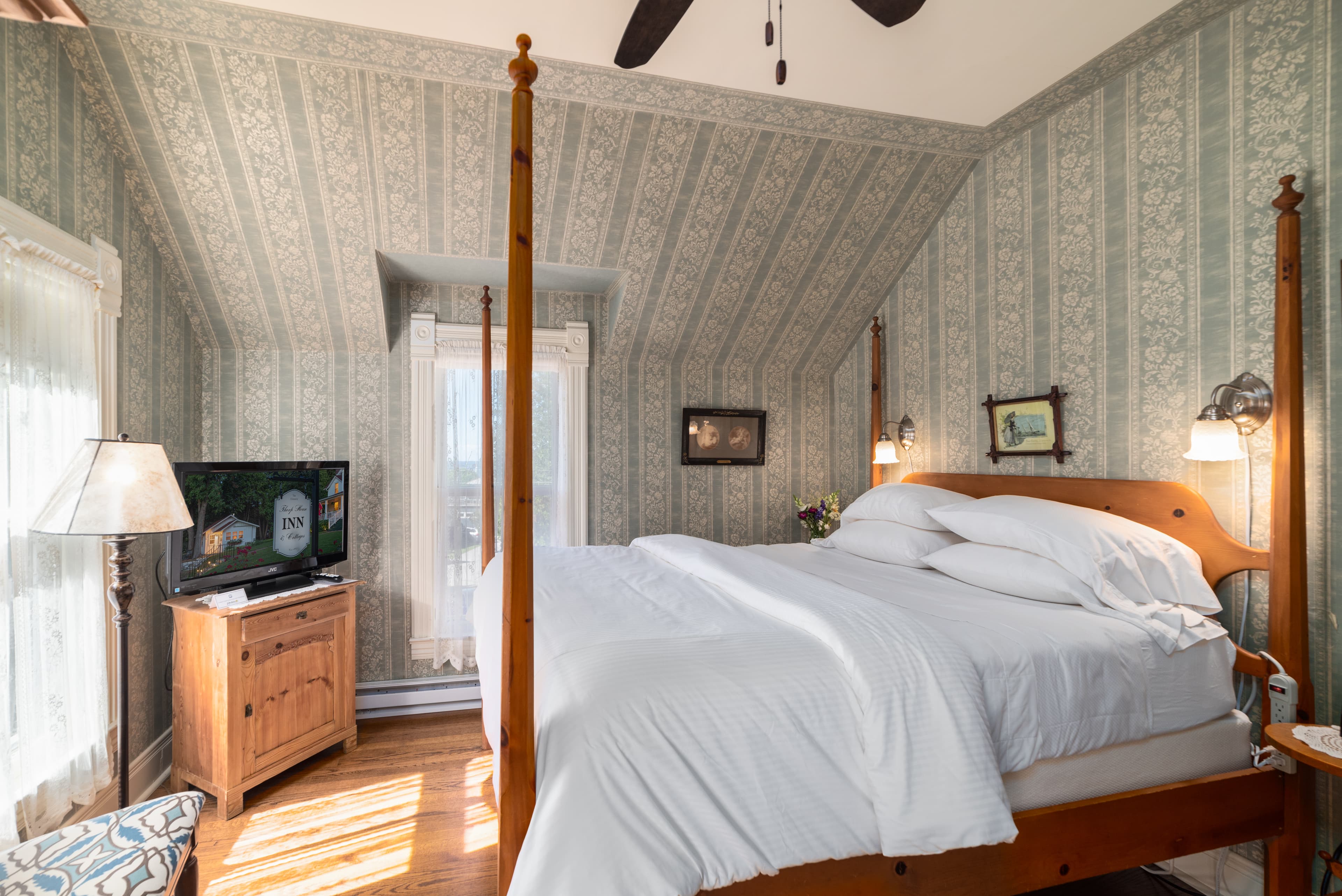 A bright bedroom with a wooden four-poster bed, patterned wallpaper, a television on a dresser, and a window letting in sunlight.