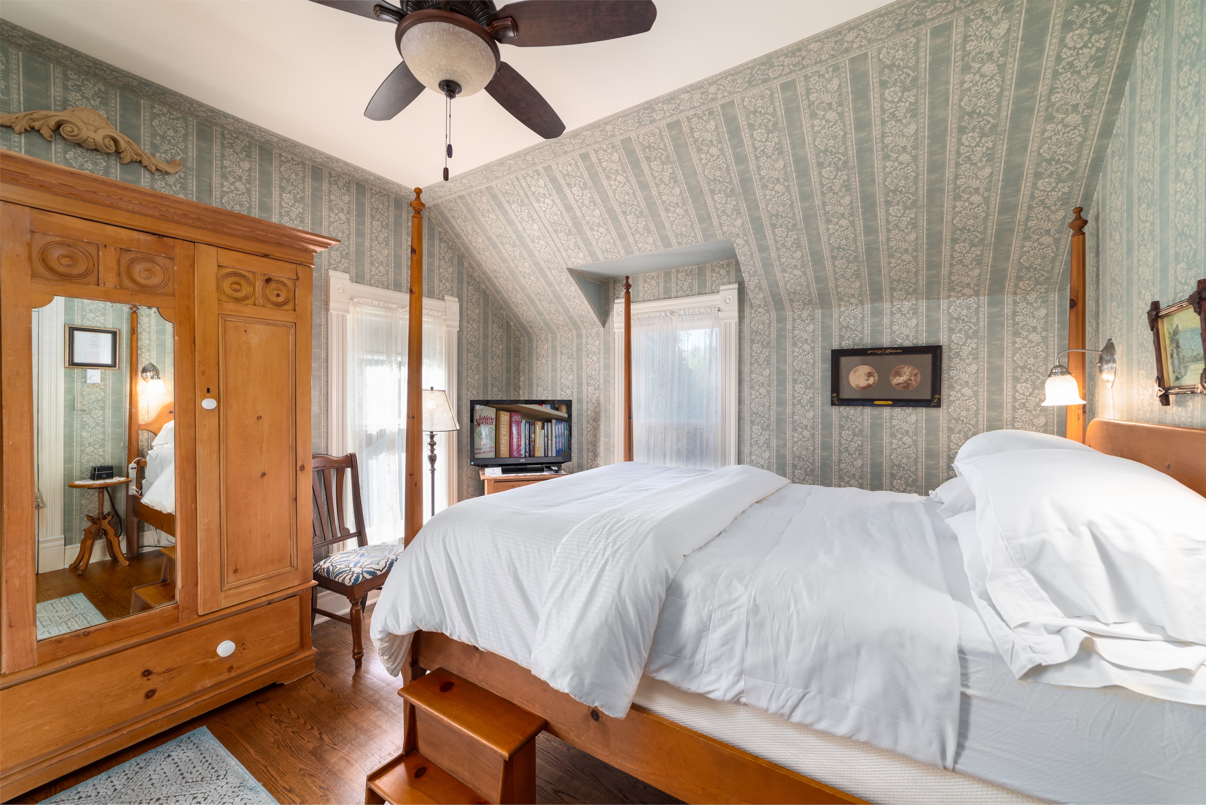 A quaint bedroom featuring a large wooden wardrobe with a mirror, a four-poster bed, and patterned wallpaper.