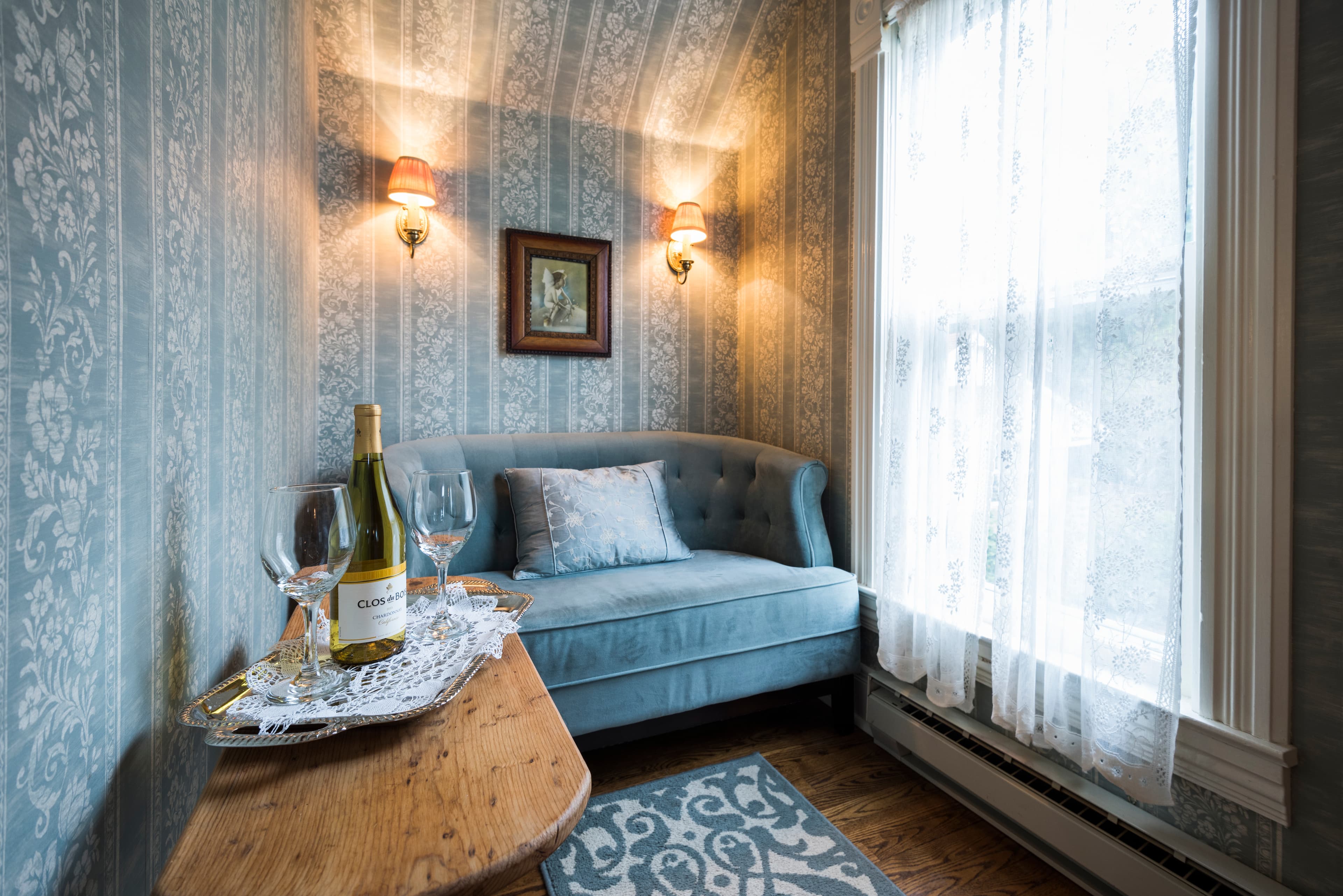 A cozy corner nook with blue patterned wallpaper, a small settee, a wooden table with wine, and a large window with sheer curtains.