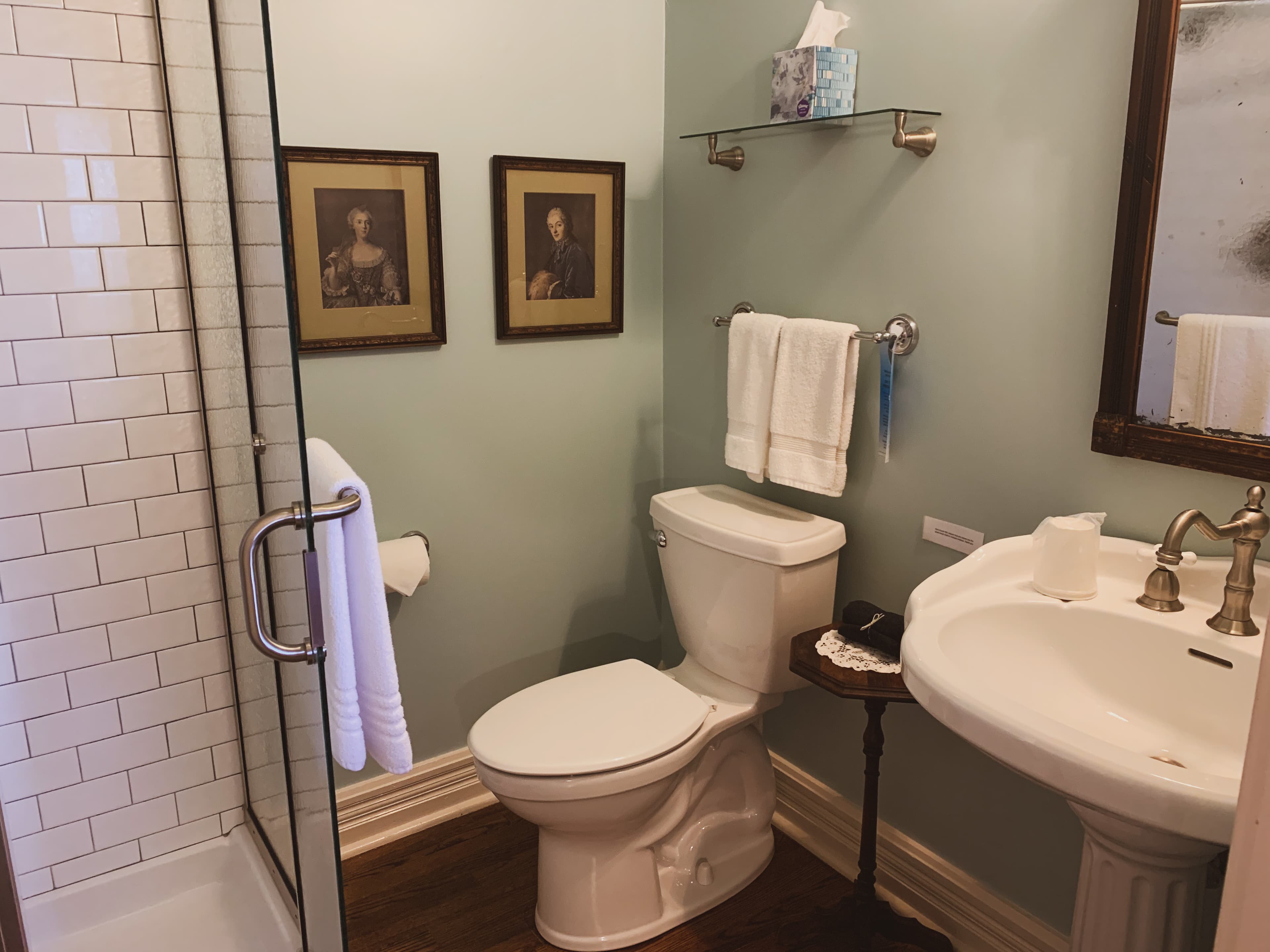 A light blue bathroom with a white pedestal sink, toilet, glass shower, and two framed pictures on the wall.