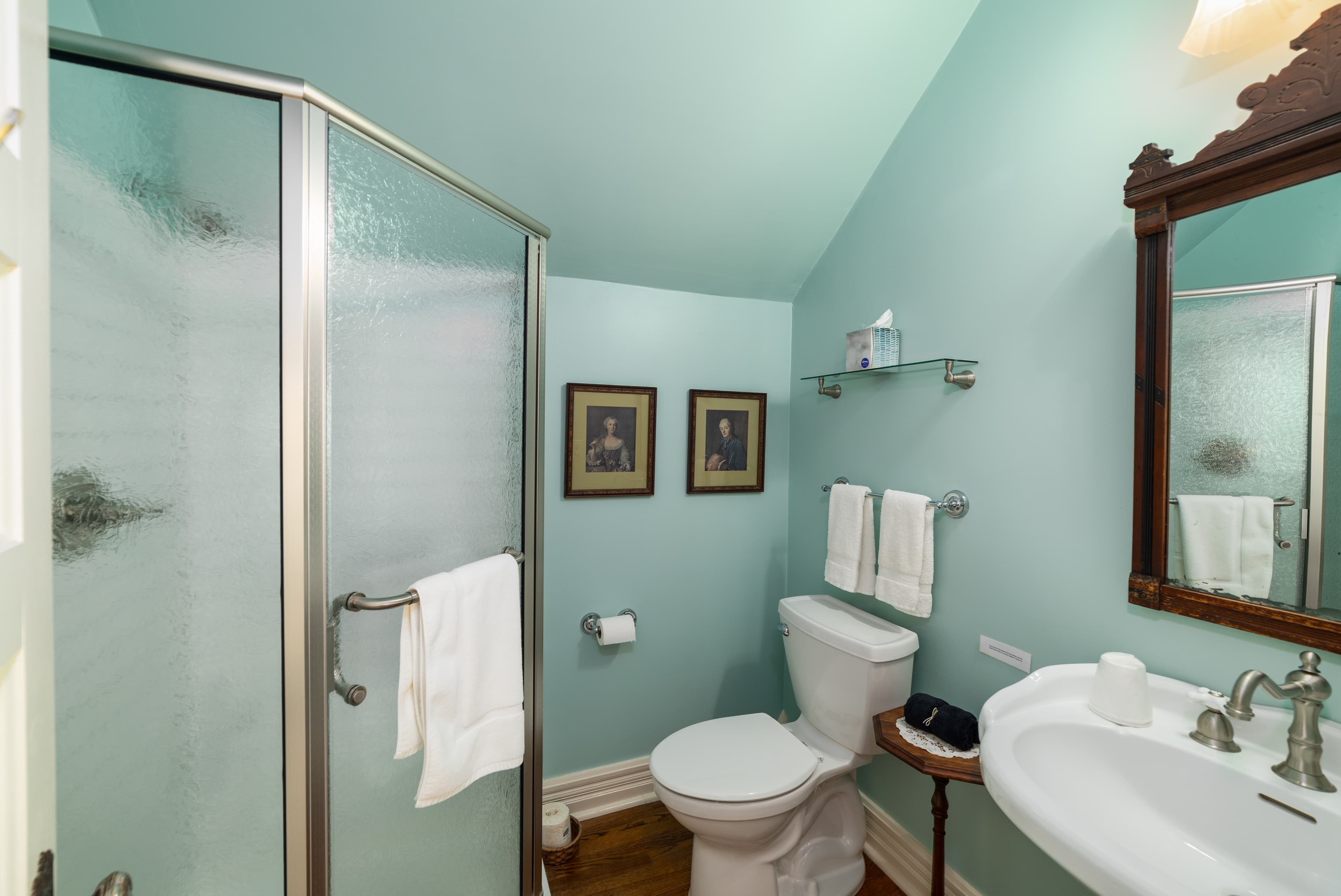 A well-appointed bathroom with light blue walls, a glass-enclosed shower, a toilet, and a sink with a dark wood framed mirror.
