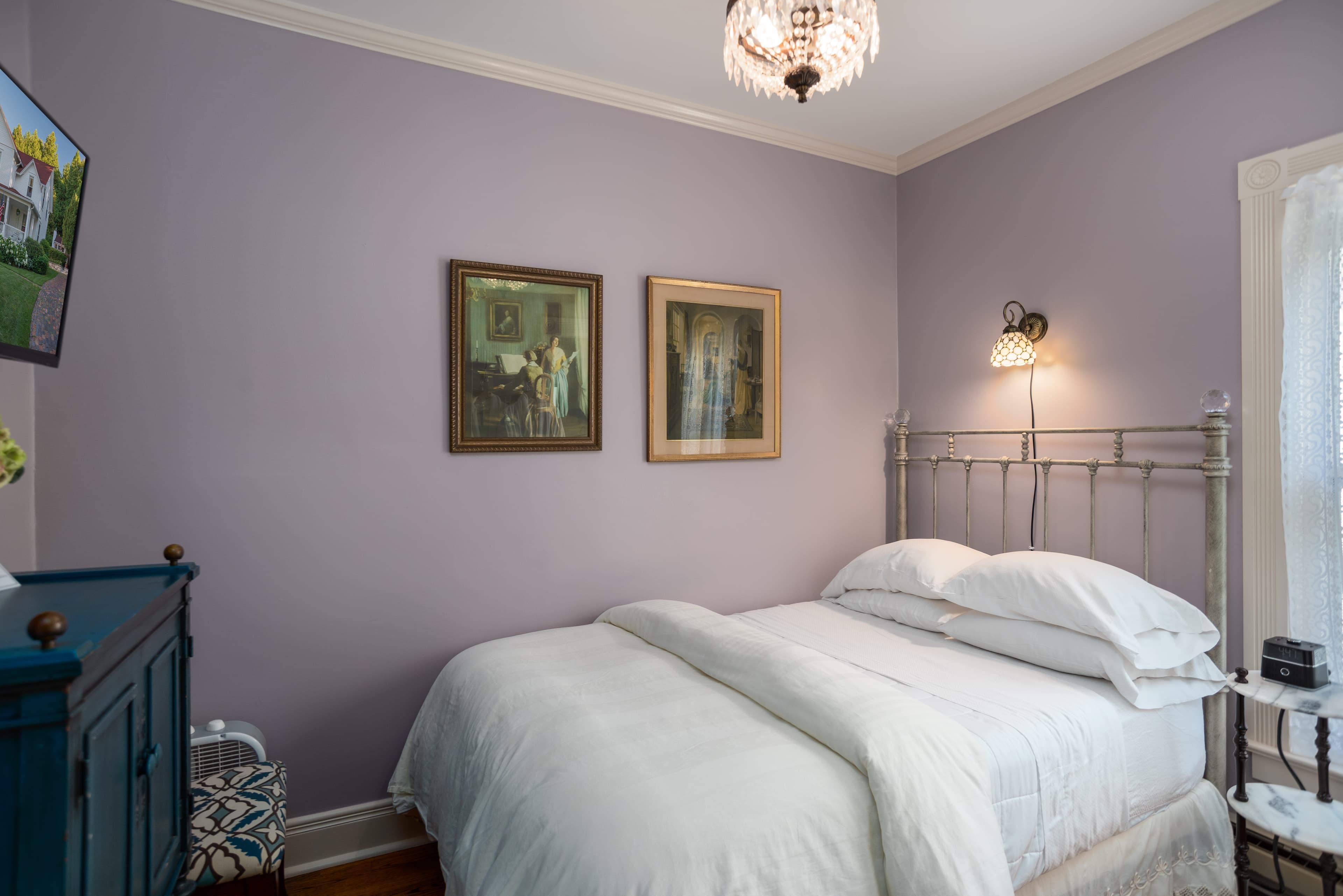 A charming bedroom featuring light purple walls, a neatly made white bed, two framed pictures, and a television on a blue dresser.