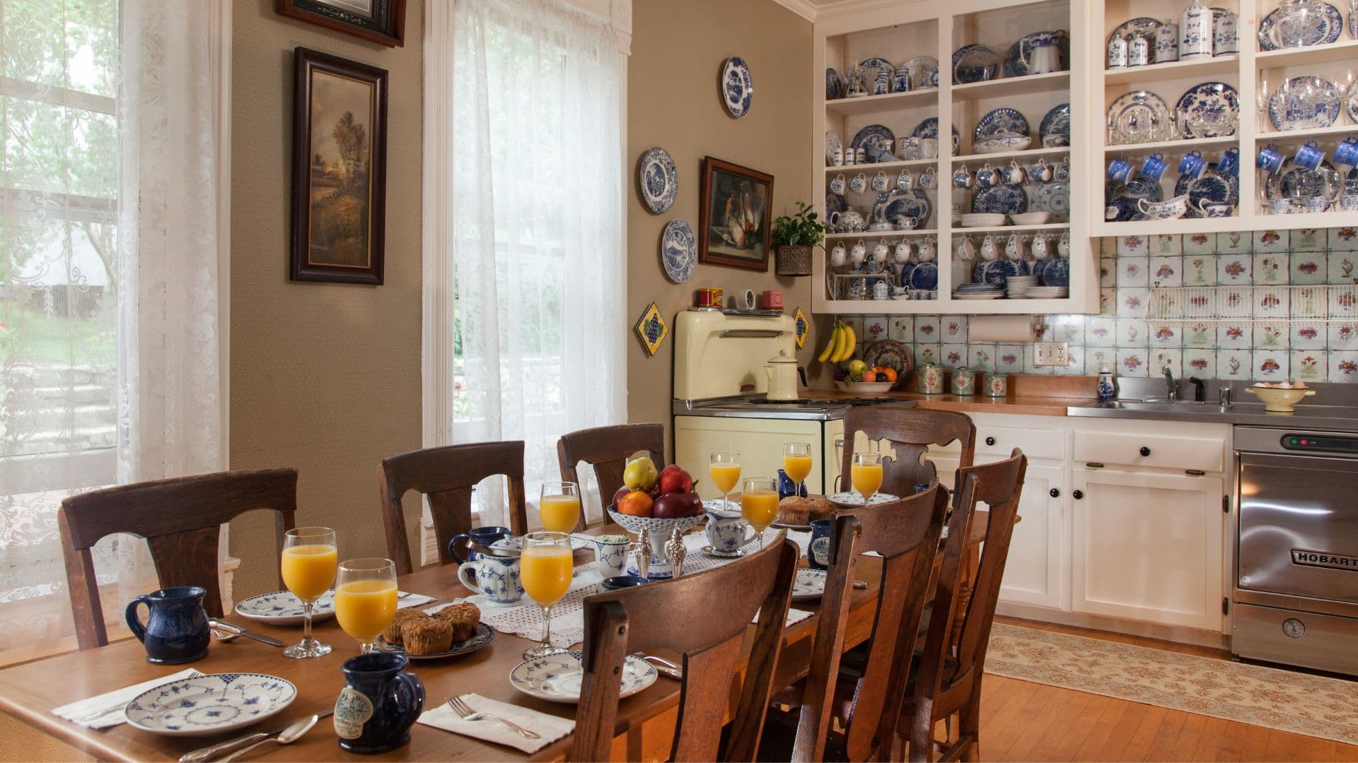 A spacious dining table set with breakfast items, including orange juice and muffins, in a cozy kitchen adorned with blue-and-white dishware.