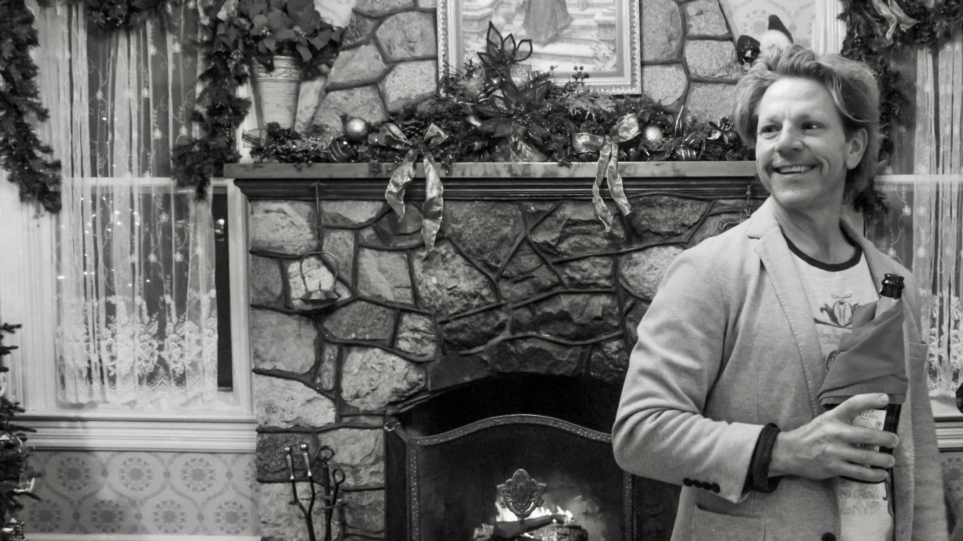 A smiling man in a blazer holds a bottle while standing in front of a stone fireplace decorated for the holidays.