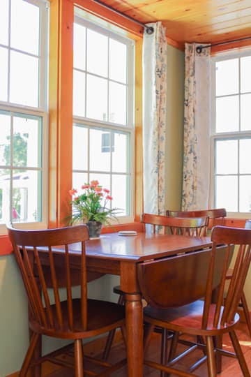 A cozy dining nook with a wooden table, four chairs, and a floral centerpiece by large windows.