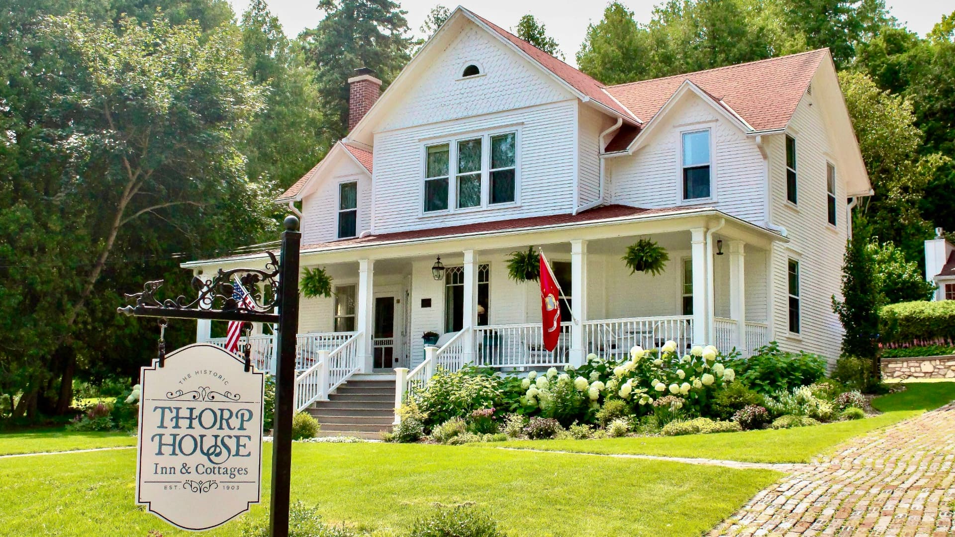 A historic white house with a porch and cottage sign, surrounded by greenery and flowers.