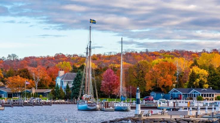 A serene harbor scene with sailboats docked, surrounded by vibrant autumn foliage and quaint buildings.