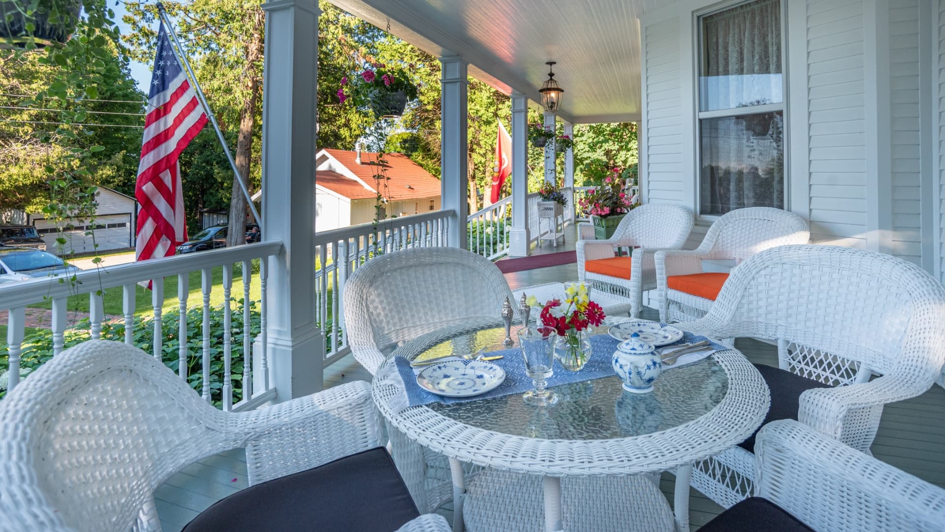 A cozy porch setting featuring white wicker furniture, a small table set for tea, and an American flag in the background.