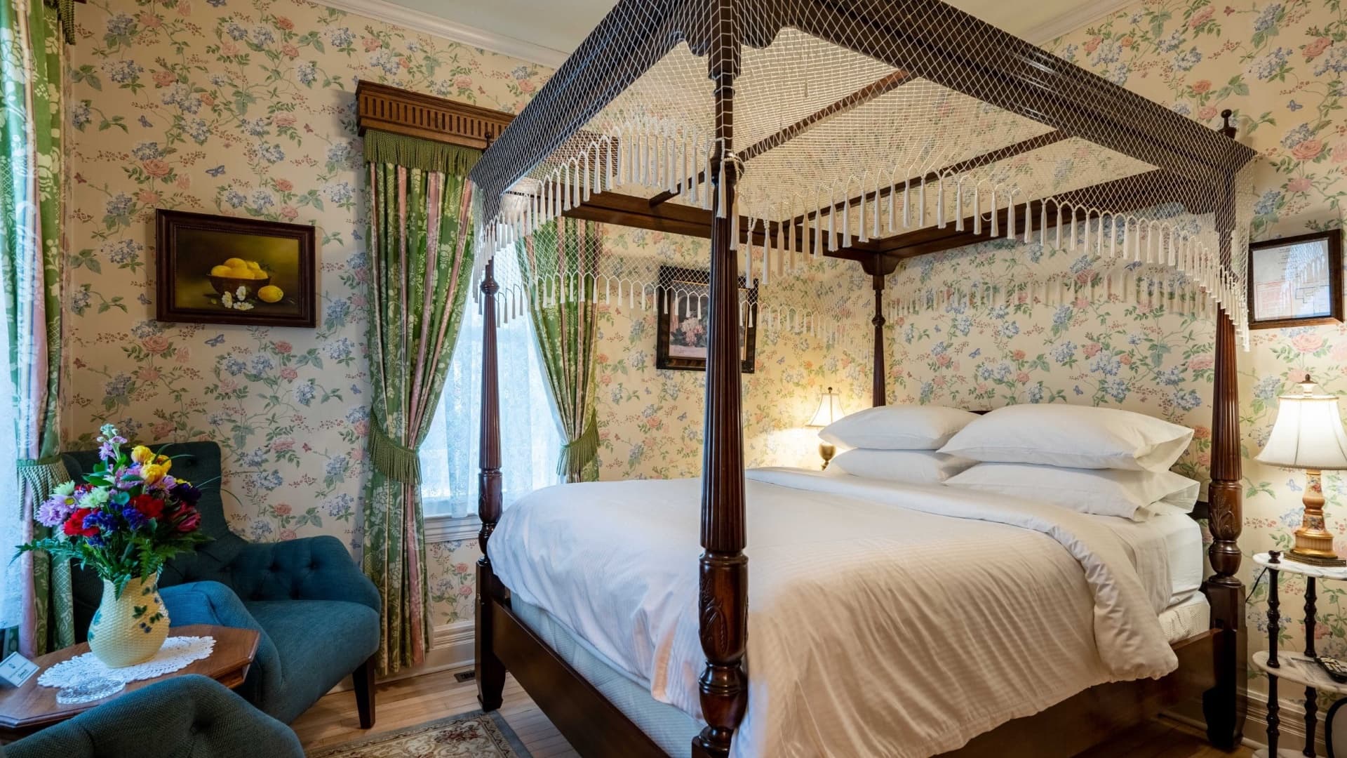 A cozy bedroom featuring a four-poster bed, floral wallpaper, and a sitting area with a vase of flowers.