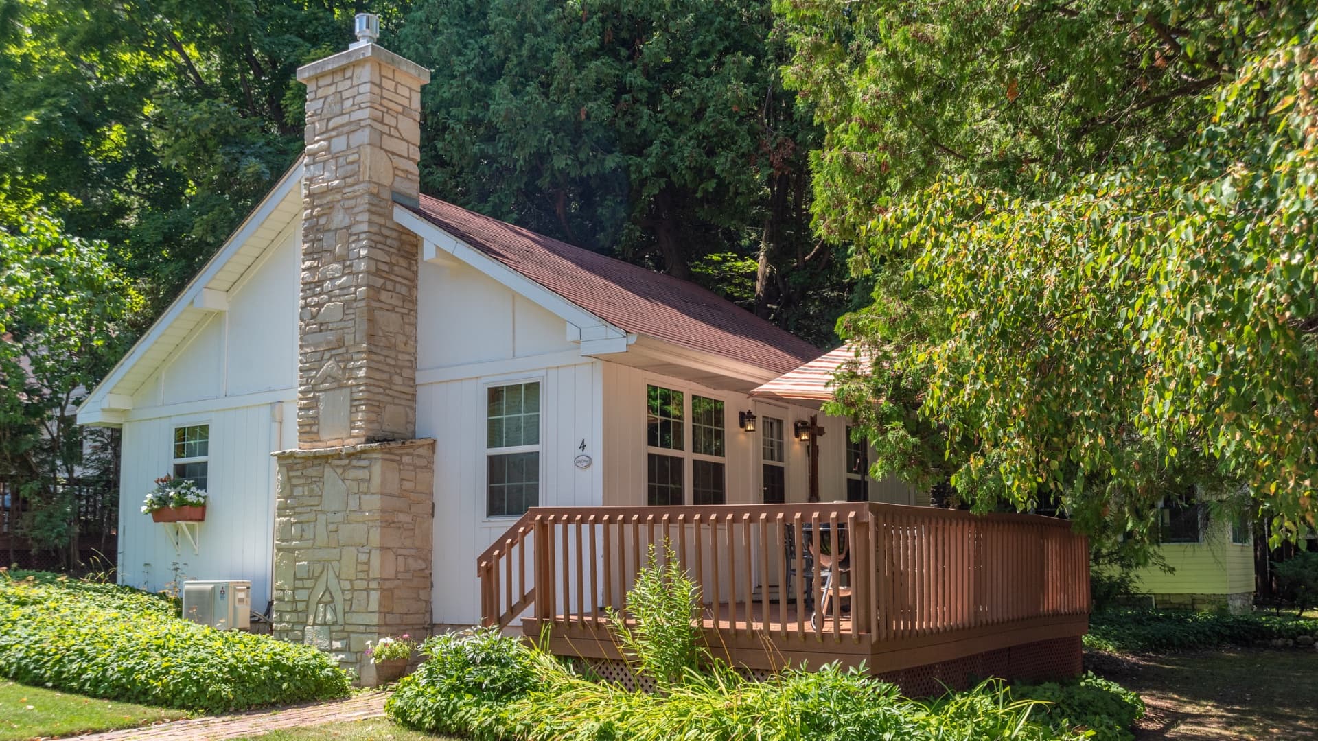 A charming white house with a stone chimney and wooden deck, surrounded by lush greenery.