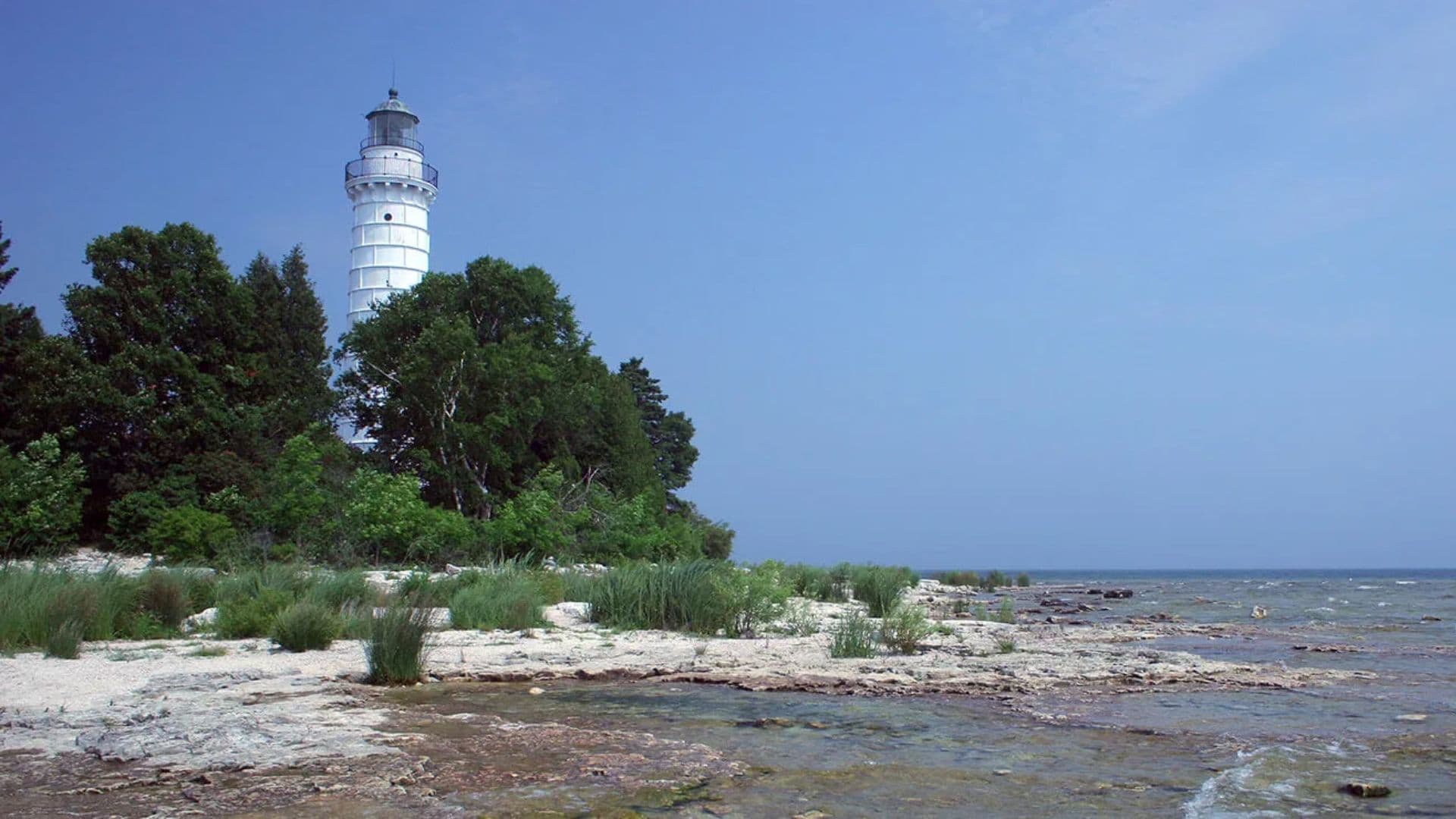 A white lighthouse stands beside a rocky shoreline surrounded by green vegetation under a clear blue sky.