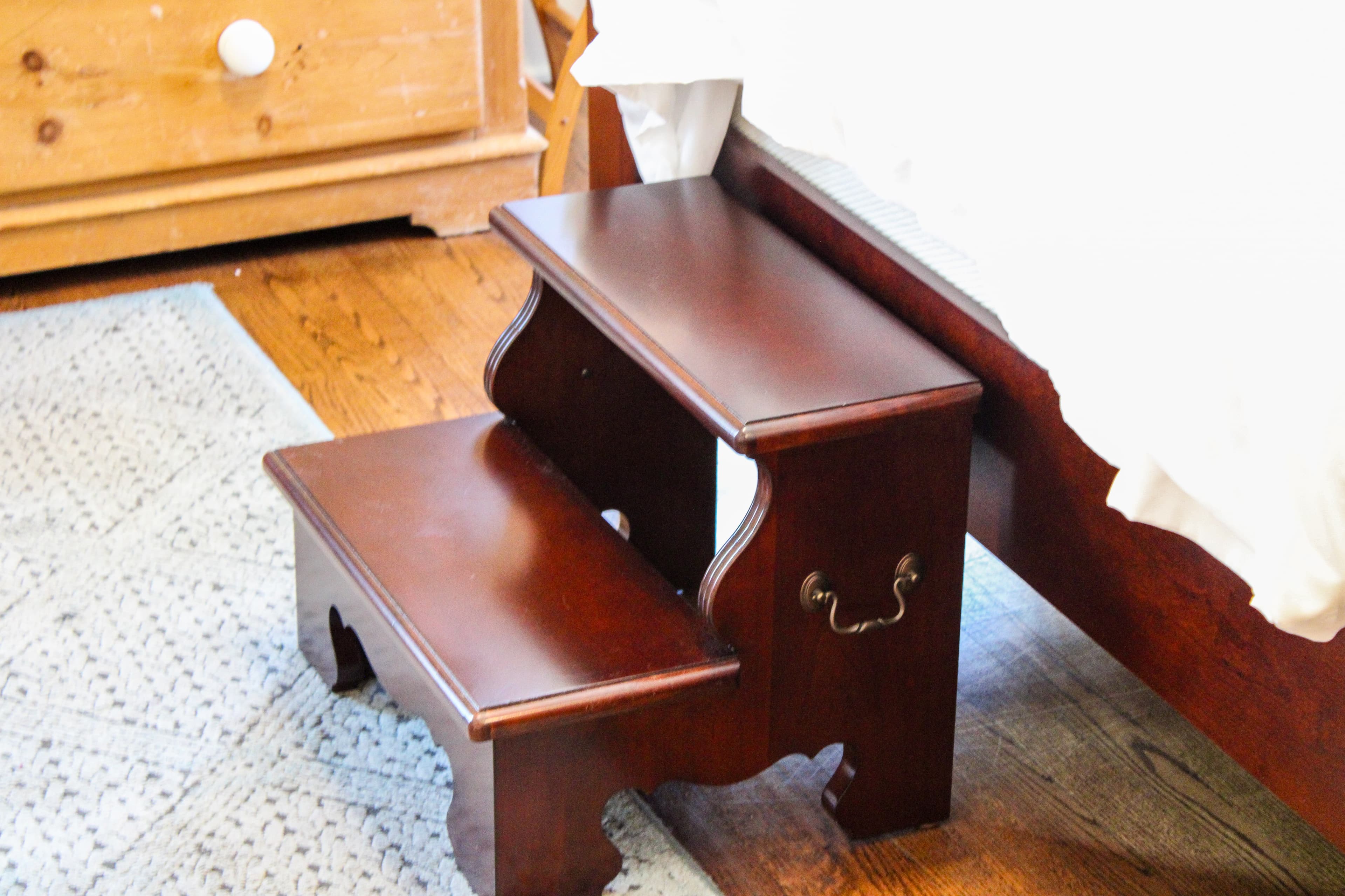A wooden two-step stool placed next to a bed on a hardwood floor.