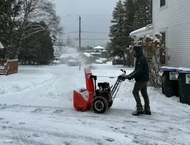 A person operates a snowblower to clear a snow-covered driveway on a winter day.