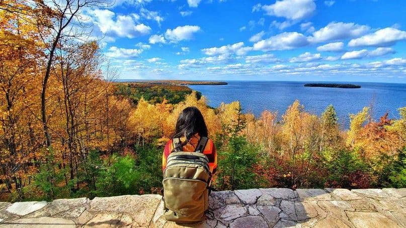 A person with a backpack gazes over a scenic autumn landscape featuring colorful trees and a calm lake.