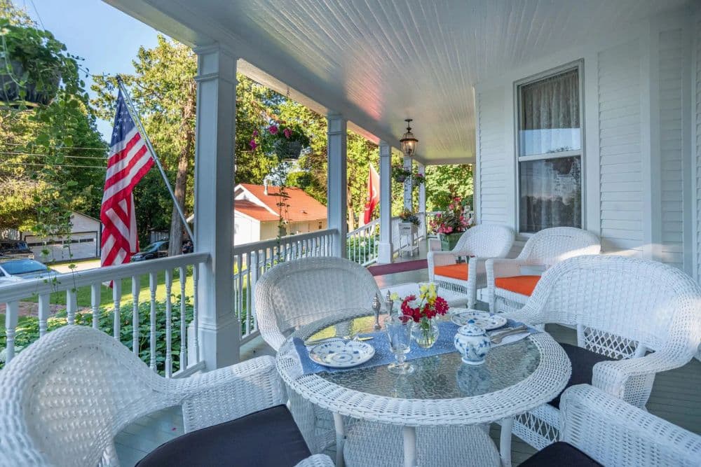 A charming porch featuring white wicker furniture, a glass table with floral arrangements, and a backdrop of greenery and an American flag.