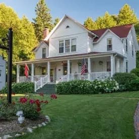 A charming two-story white house with a red roof, set amidst lush greenery and flower beds.
