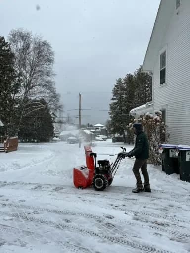 A person using a red snowblower machine on the driveway outside a home